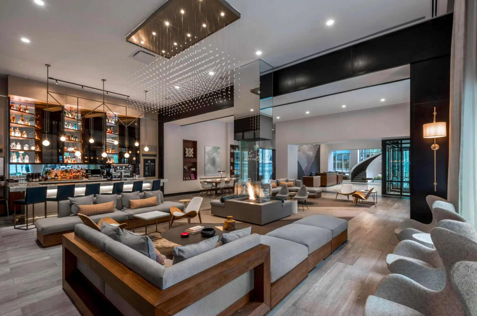 Modern hotel lobby with a large seating area including sofas and armchairs, a bar with illuminated bottles, a coffee table, a ceiling chandelier, a fireplace, and contemporary decor in neutral tones.