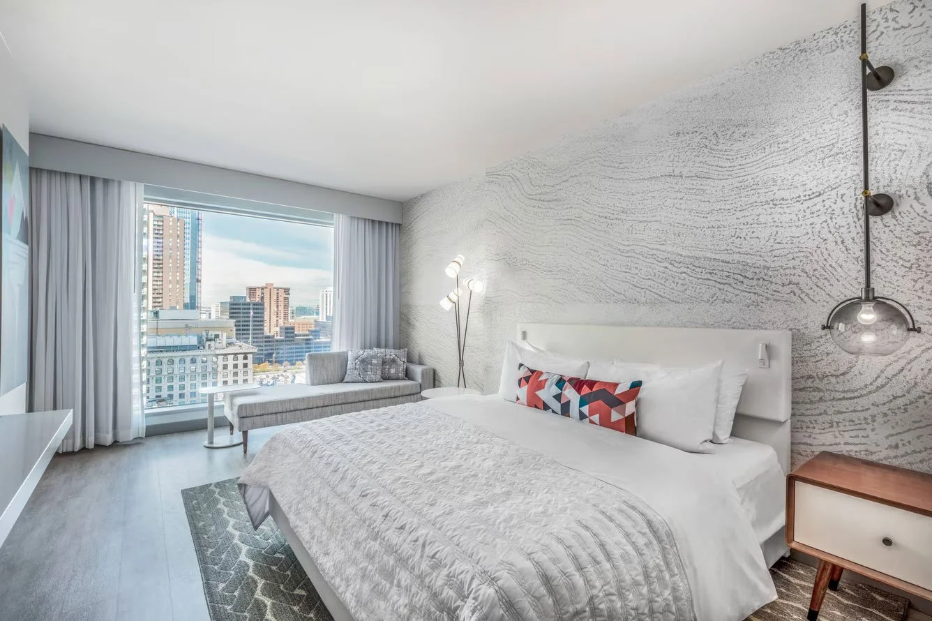 Modern bedroom with a large window view of city buildings, white bed with decorative pillows, bedside table, floor lamp, and wall-mounted pendant light.