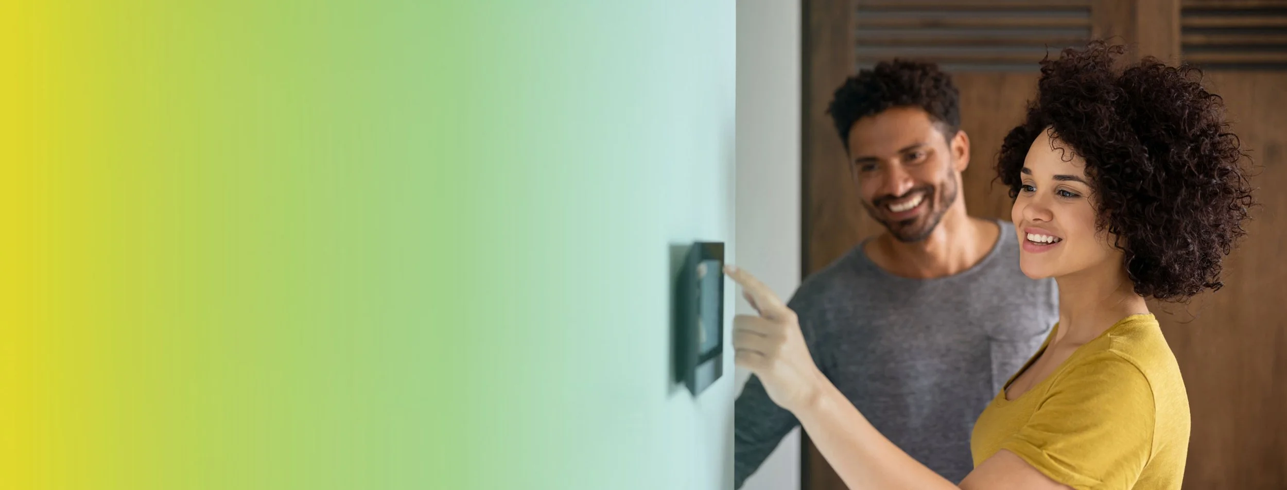 A woman in a yellow shirt smiling as she presses a button on a smart home control panel on a green wall, with a man in a gray shirt standing next to her, smiling and observing.