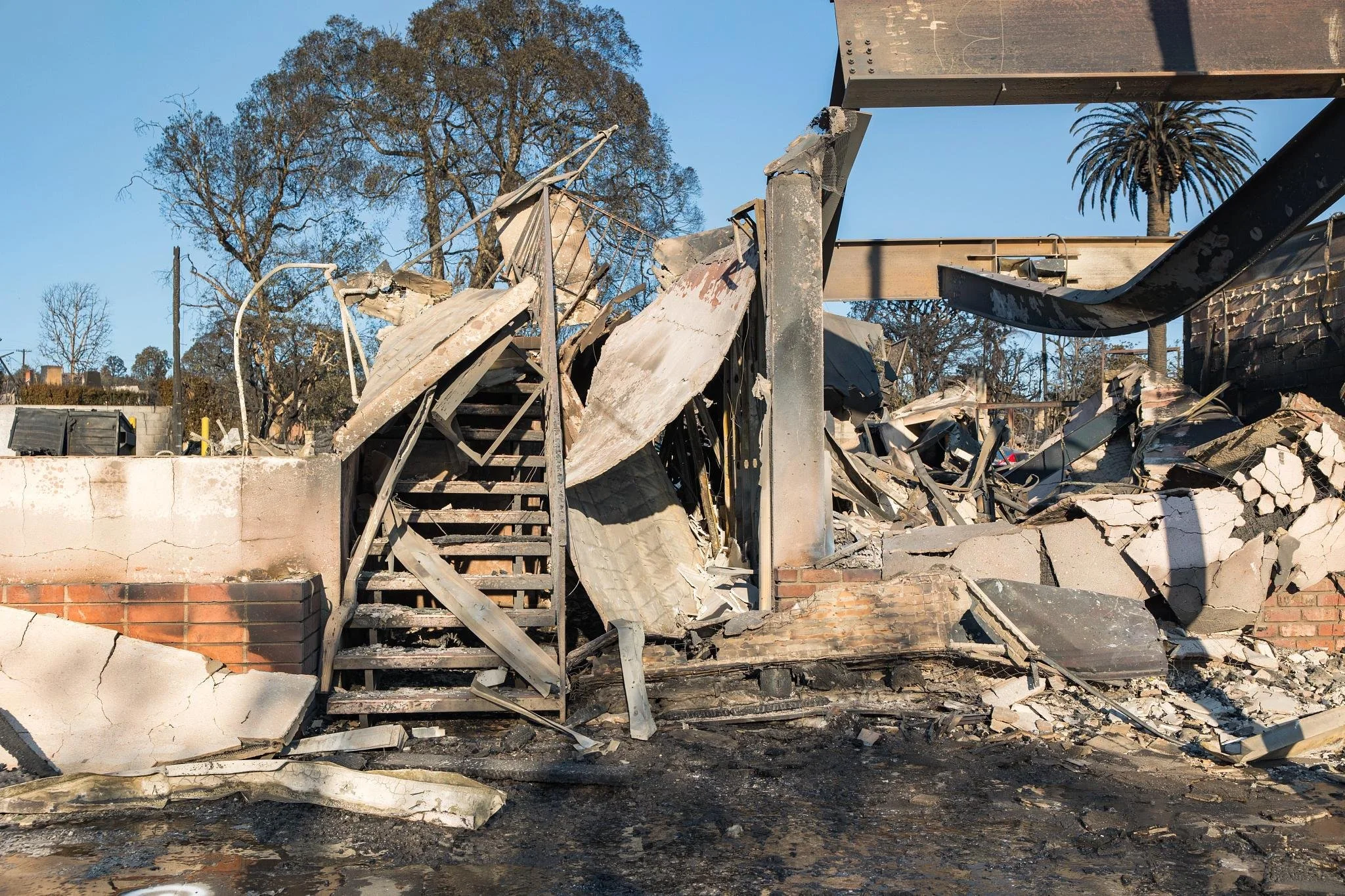 Ruined building with collapsed walls and debris, charred remains, and torn roofing material, with trees in the background and a clear blue sky.