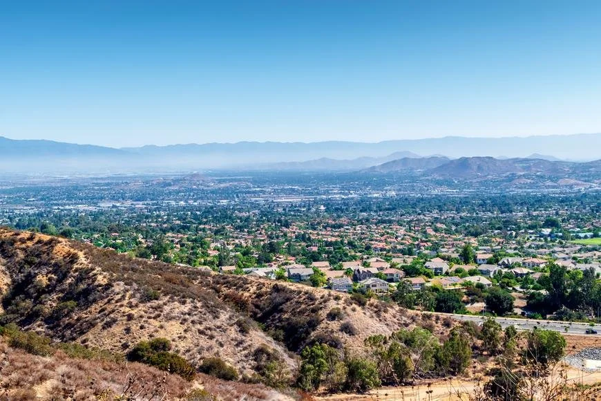 A panoramic view of a city from a hillside, with residential neighborhoods, green trees, and distant mountains under a clear blue sky.