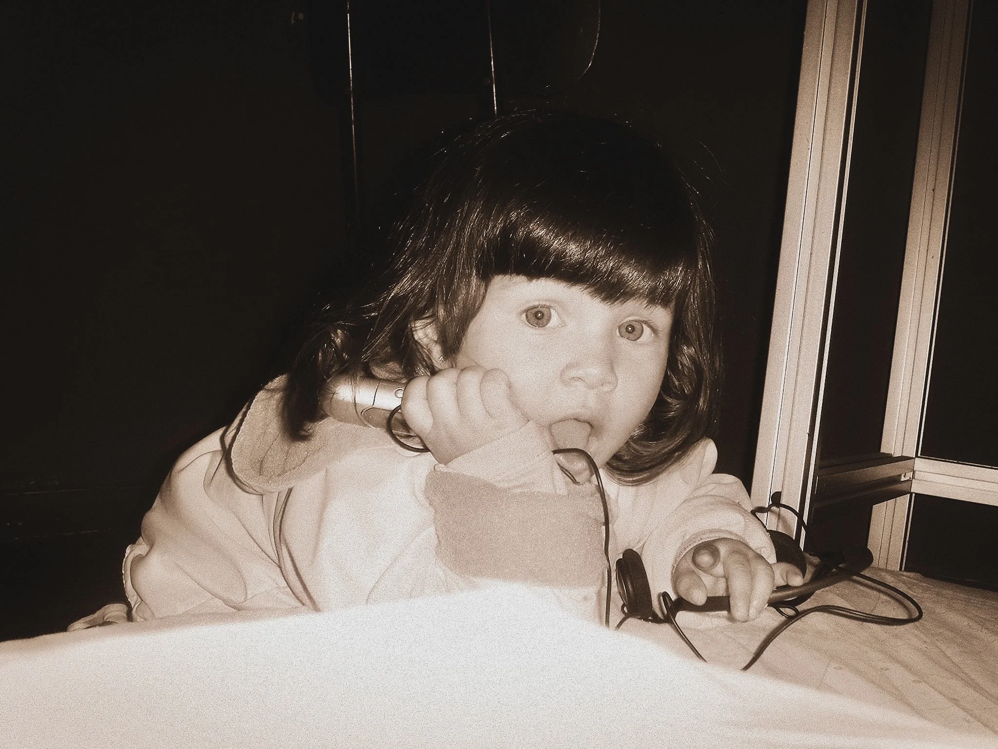 A young girl with curly dark hair and light skin resting her chin on her hand, sitting at a table with electronic device and wires. Background includes a metallic or glass cabinet.