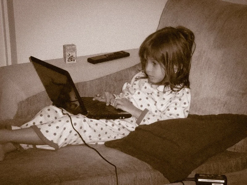 A young girl in polka dot pajamas lying on a couch, watching or using a laptop. There is a remote control and a small box on the wall behind her.