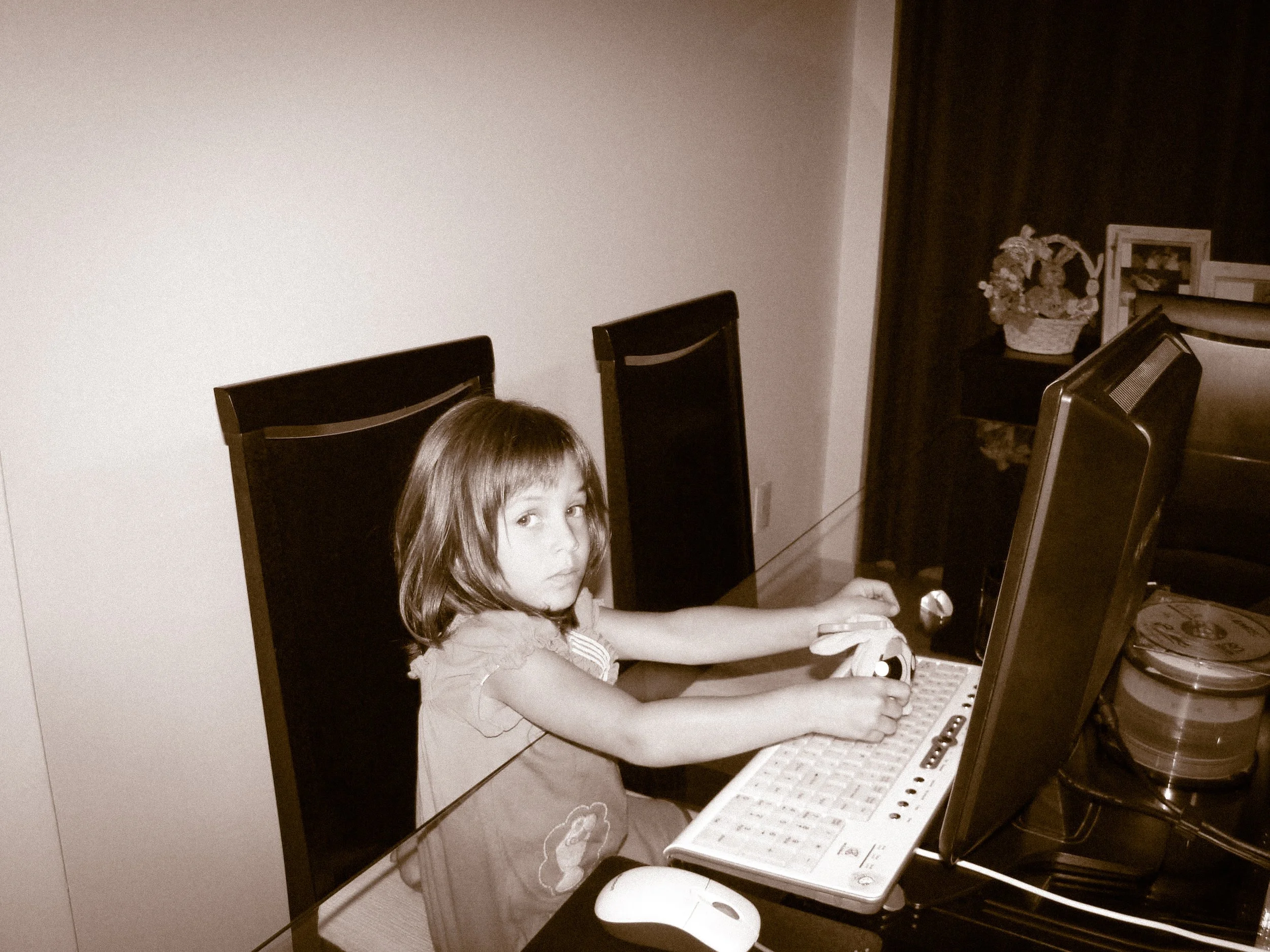 A young girl with long brown hair sitting at a kitchen table, looking at a laptop computer. The table has a keyboard and a computer mouse, and there are stacked containers and decorative items on a side table in the background.