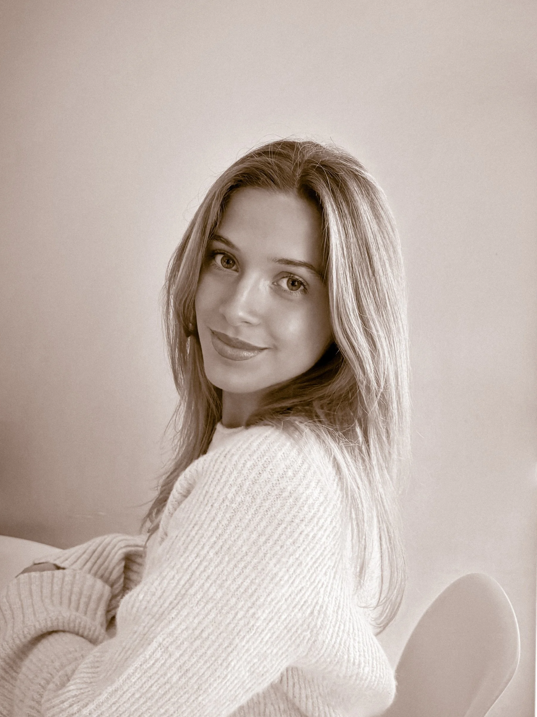 A woman with long hair smiling at the camera, wearing a white sweater, in a minimalist room.