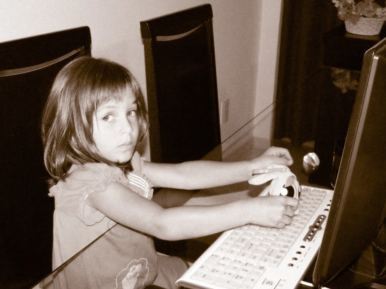 A young girl with shoulder-length hair sitting at a desk, using a desktop computer with an old keyboard and a mouse, looking at the camera in a sepia photo.