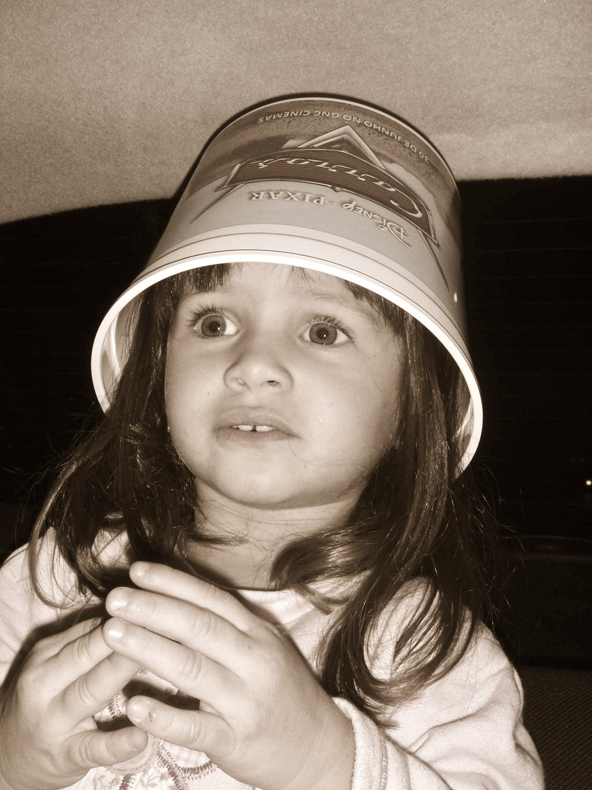 A young girl with long hair, wide eyes, and a surprised expression, looking through the top of a Cars movie popcorn bucket.