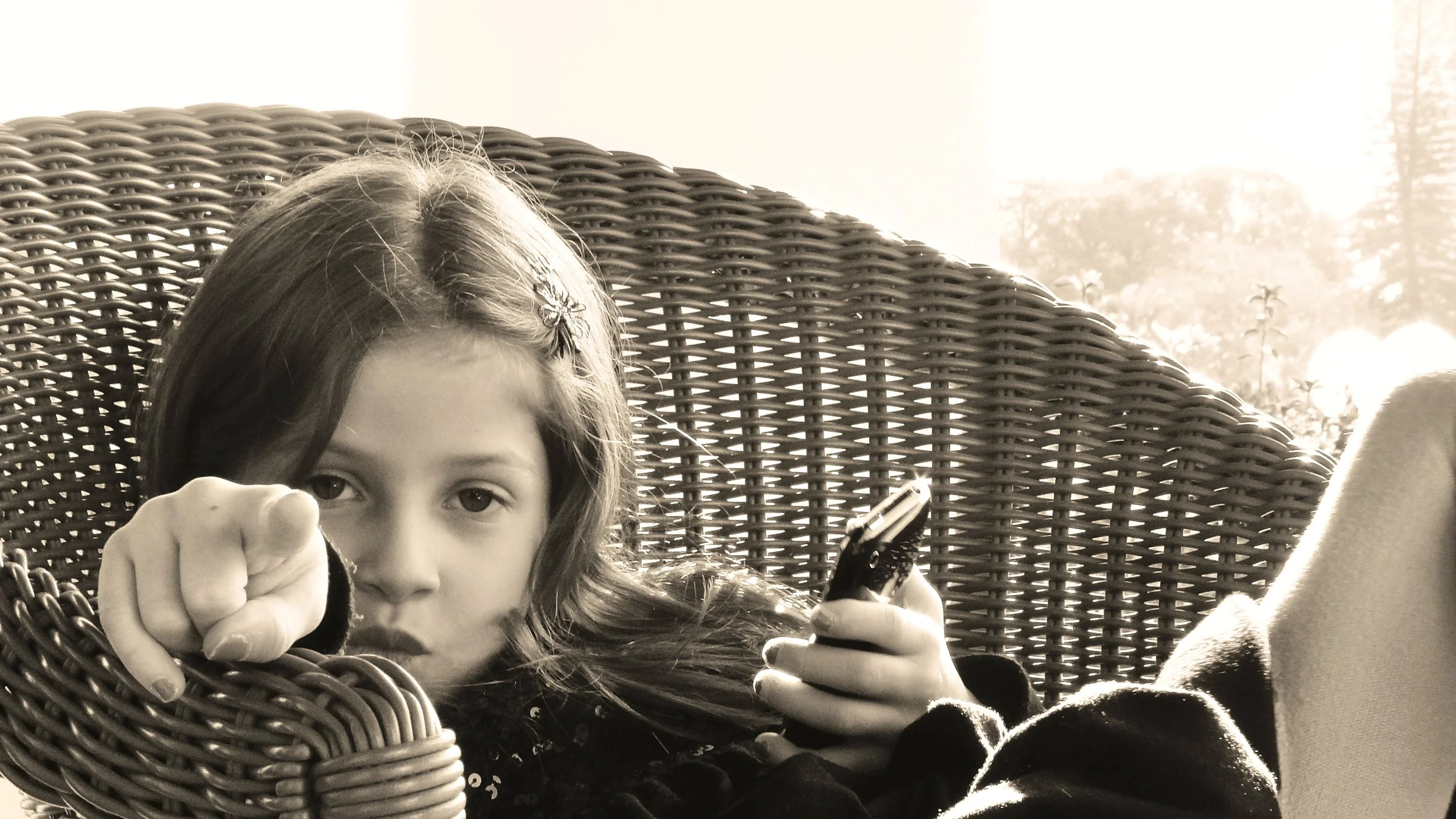 A young girl with long hair and a flower hairpin, sitting on a wicker chair outdoors, holding a phone and pointing at the camera.