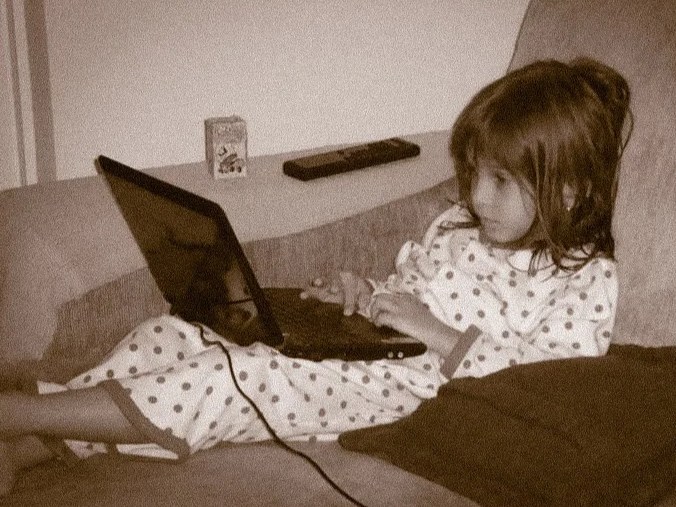 A young girl in pajamas sitting on a couch, using a laptop.