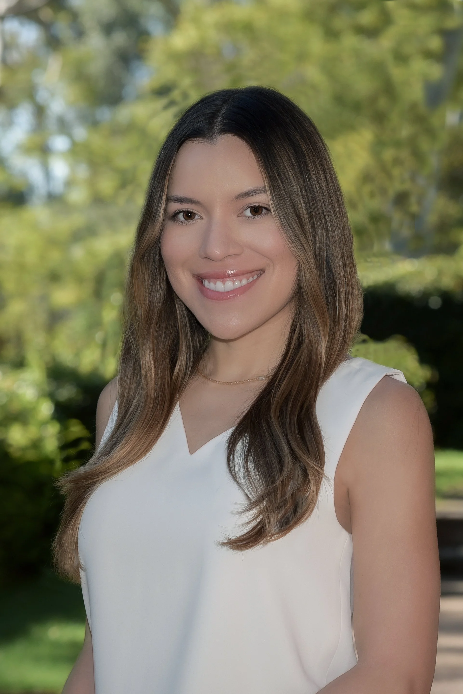 Young woman with long wavy brown hair, wearing a white sleeveless top, smiling outdoors with green trees in the background.