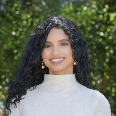 A young woman with curly black hair, smiling, wearing a light gray top and earrings, with a blurred green foliage background.