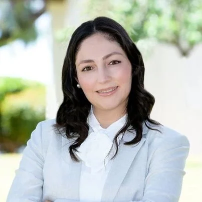 Portrait of a smiling woman with dark hair wearing a light blue blazer and white blouse, standing outdoors with green trees in the background.