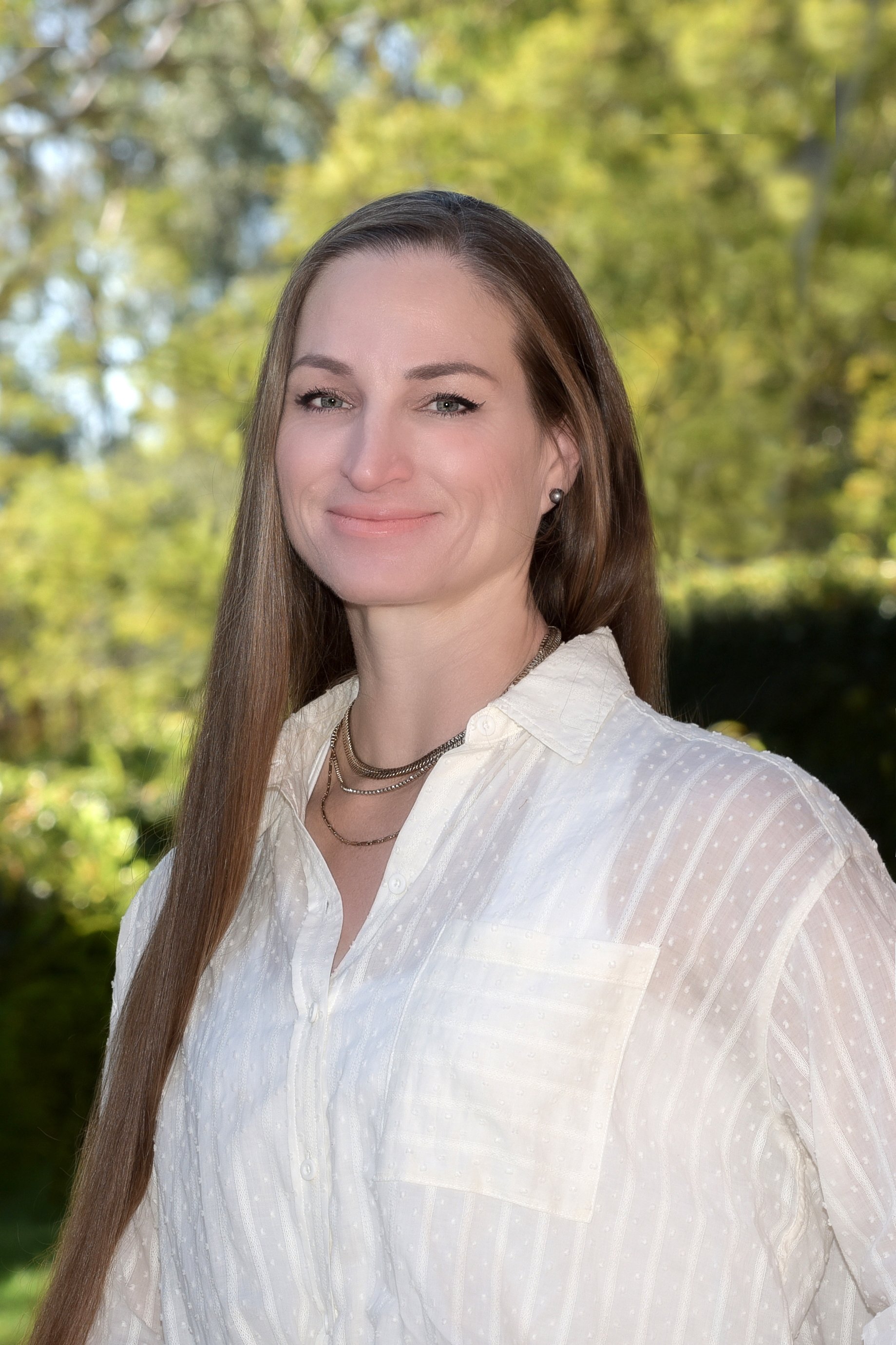 A woman with long brown hair wearing a white blouse and layered necklaces outdoors, with a background of trees with green leaves.