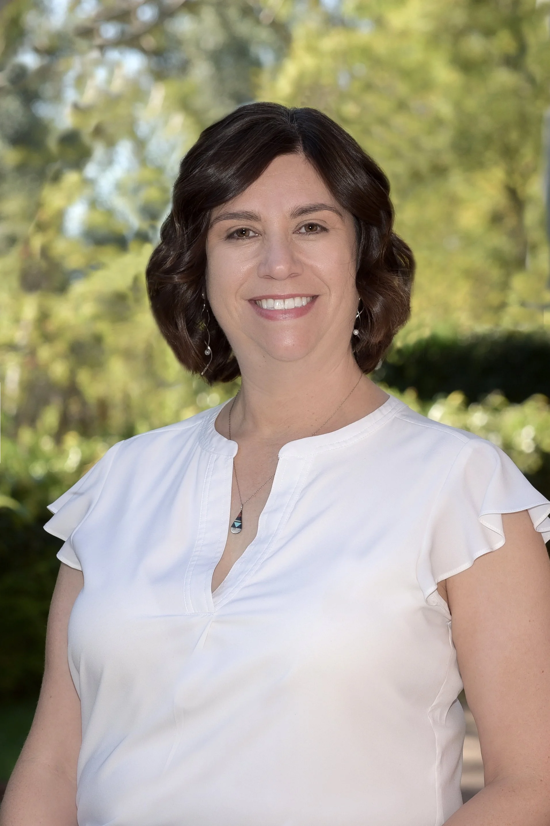 A woman with dark brown hair smiling outdoors with green trees in the background, wearing a white top and earrings.
