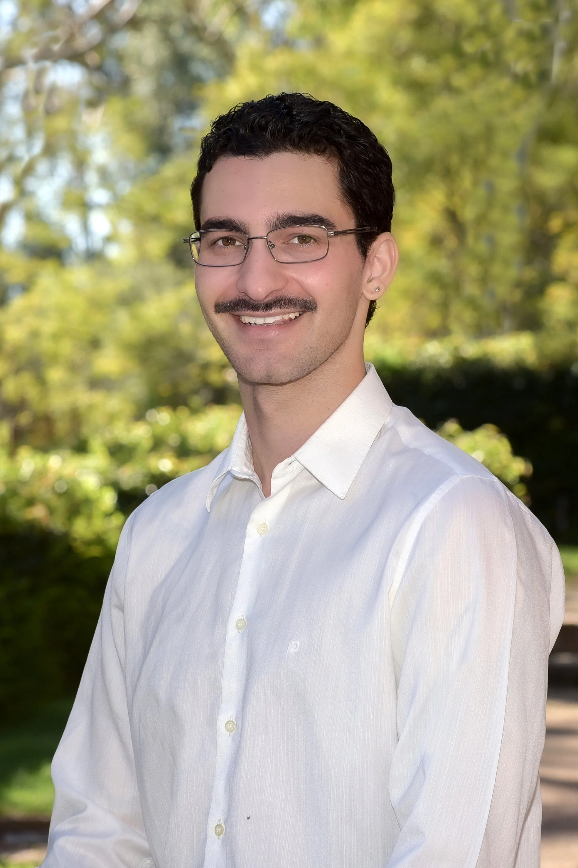 A man with short dark curly hair, glasses, and a mustache smiling, wearing a white dress shirt, outdoors with blurred green and yellow foliage in the background.