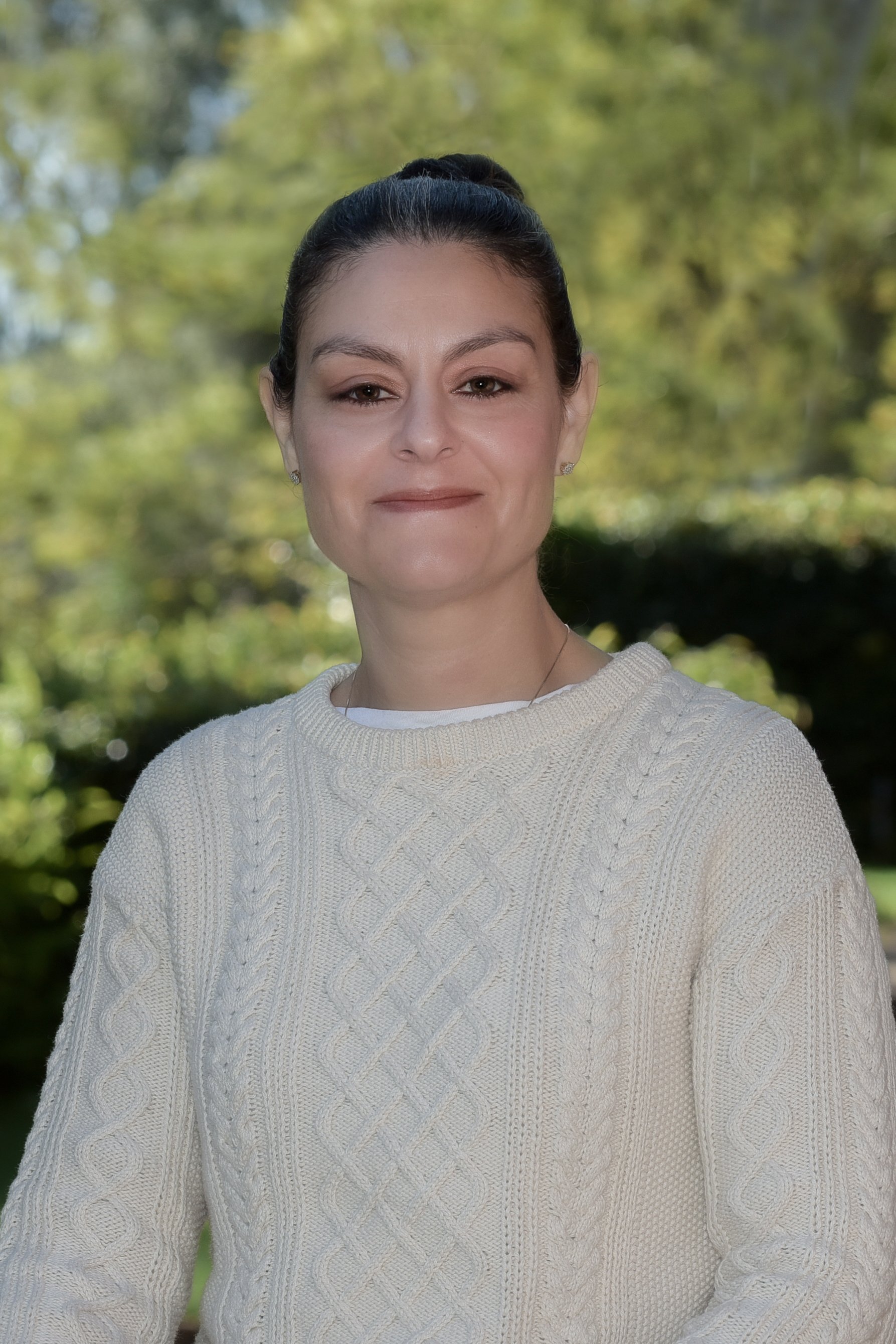 A woman with dark hair tied back, wearing a white cable knit sweater, standing outdoors with blurred green trees in the background.