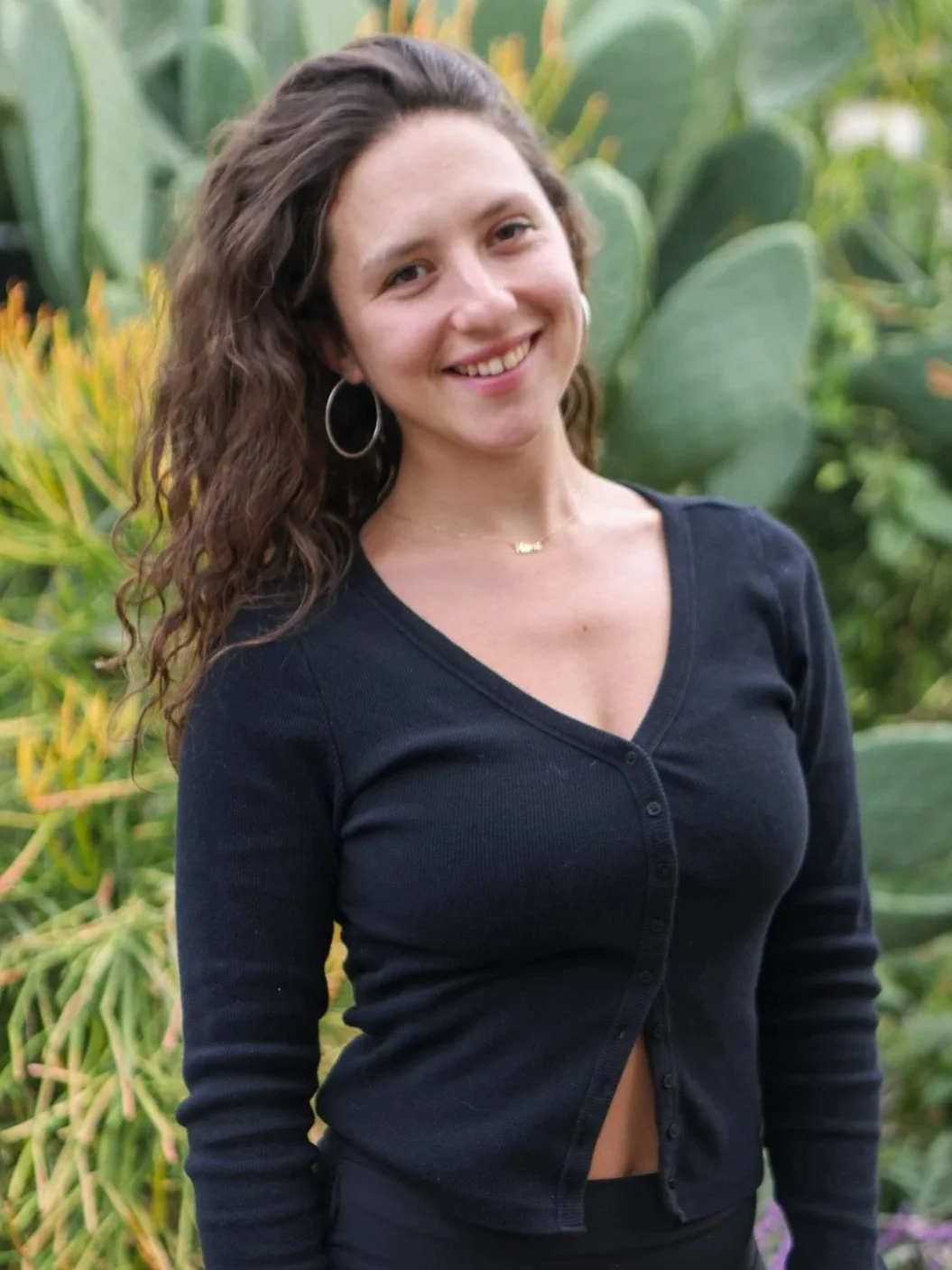 A young woman smiling outdoors with green plants in the background. She has long, curly brown hair, hoop earrings, and is wearing a black button-up shirt with a v-neck.