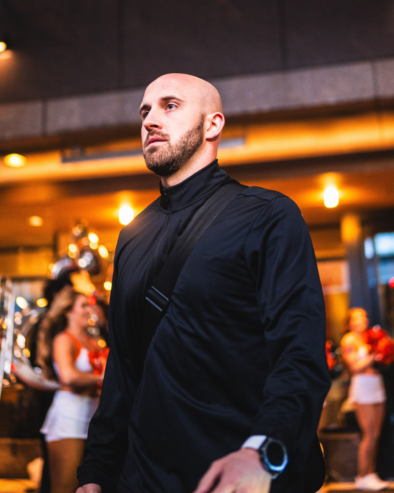 A bald man with a beard, wearing a black jacket and a smartwatch, standing in a lively indoor setting with warm lighting, with several women in cheerleader-like outfits in the background.