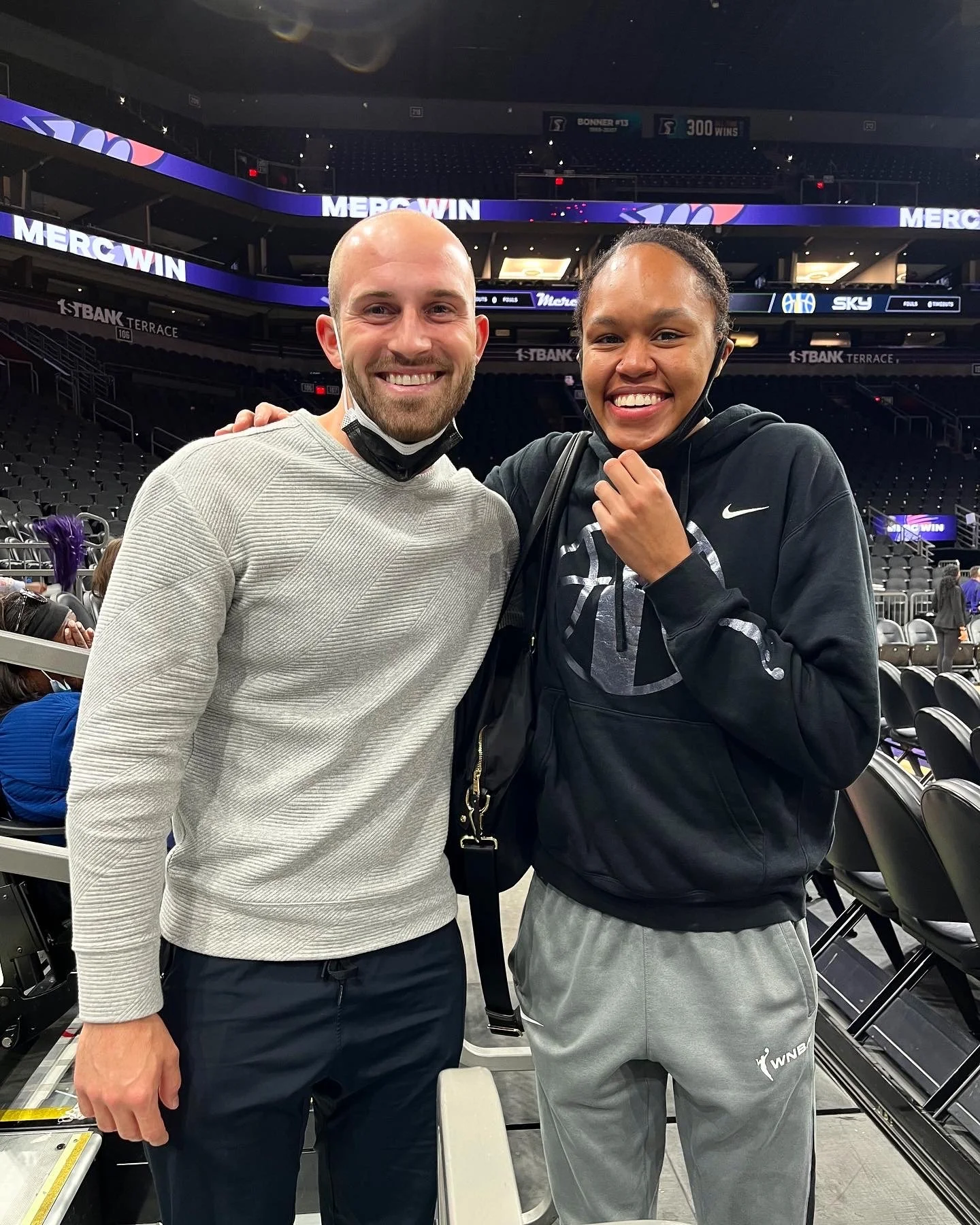 Two smiling people standing close together in an indoor sports arena, with stadium seating and electronic scoreboards in the background. One is a bald man wearing a light gray long sleeve shirt and dark pants, the other is a woman with braided hair wearing a black Nike hoodie and gray sweatpants, both holding face masks.