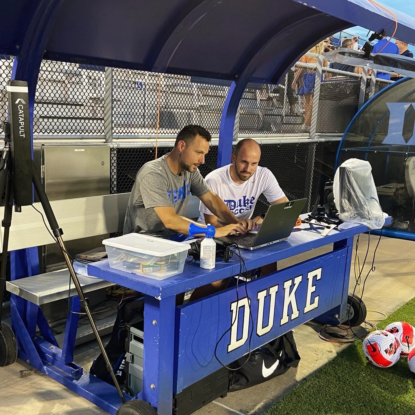 Two men working at a blue Duke University sports bench under a blue canopy, in a sports stadium. One is wearing a gray shirt and the other a white Duke shirt, both focused on a laptop. Soccer balls are on the ground nearby, and behind them is a metal fence with spectators in the background.