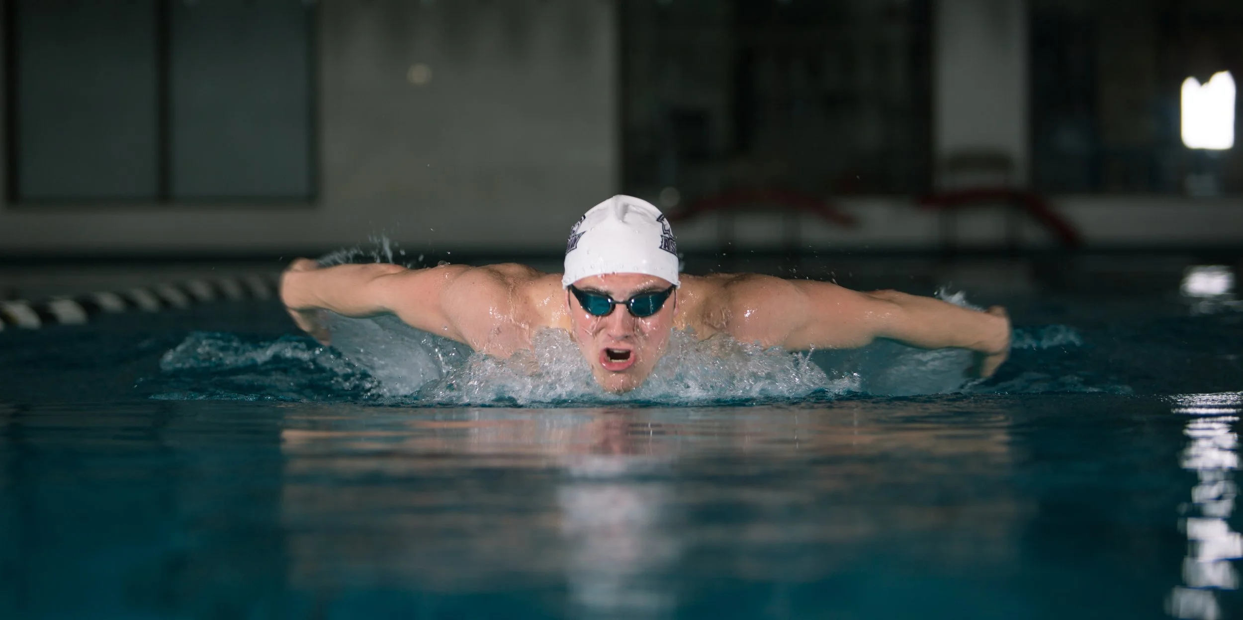 A male swimmer in a competitive swimming pool performing the butterfly stroke, wearing goggles, a swim cap, and actively propelling himself through the water with arms extended.