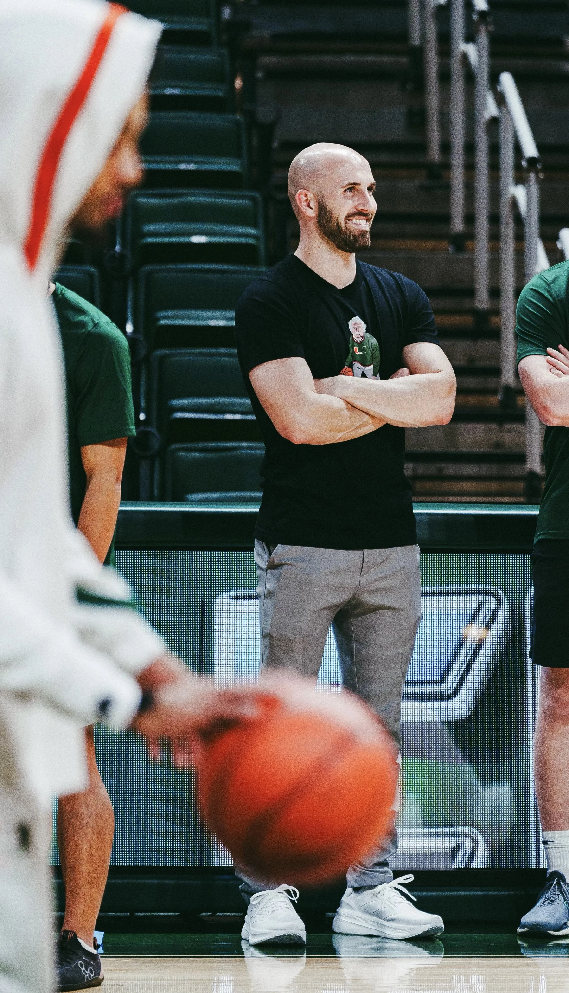 A basketball coach standing with arms crossed during a game or practice in an indoor gym, surrounded by players. The coach is smiling, has a shaved head, beard, and is wearing a black t-shirt with a cartoon character and gray pants.