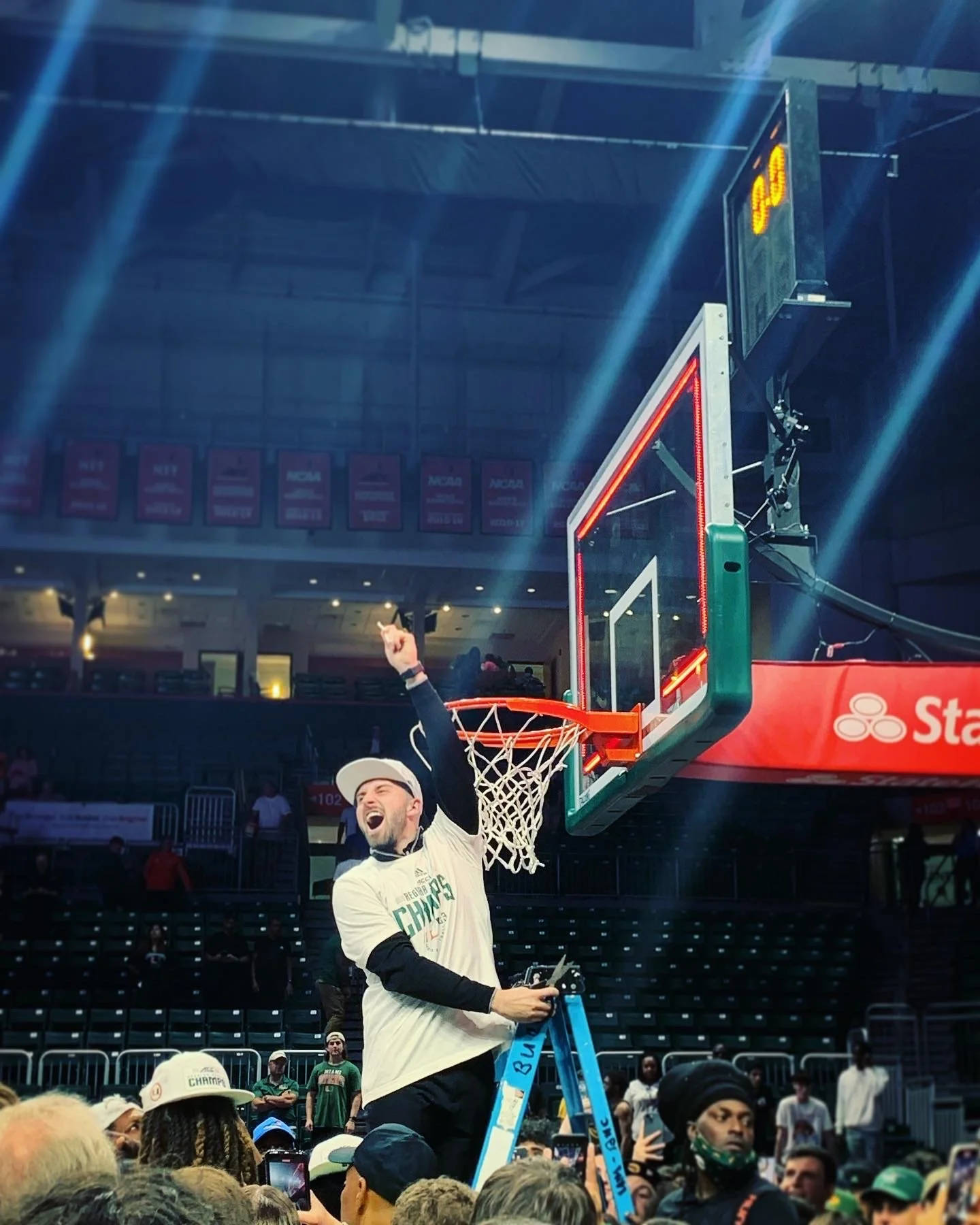 A man celebrating after winning a basketball championship, standing on a ladder near a basketball hoop with a crowd watching in the background.