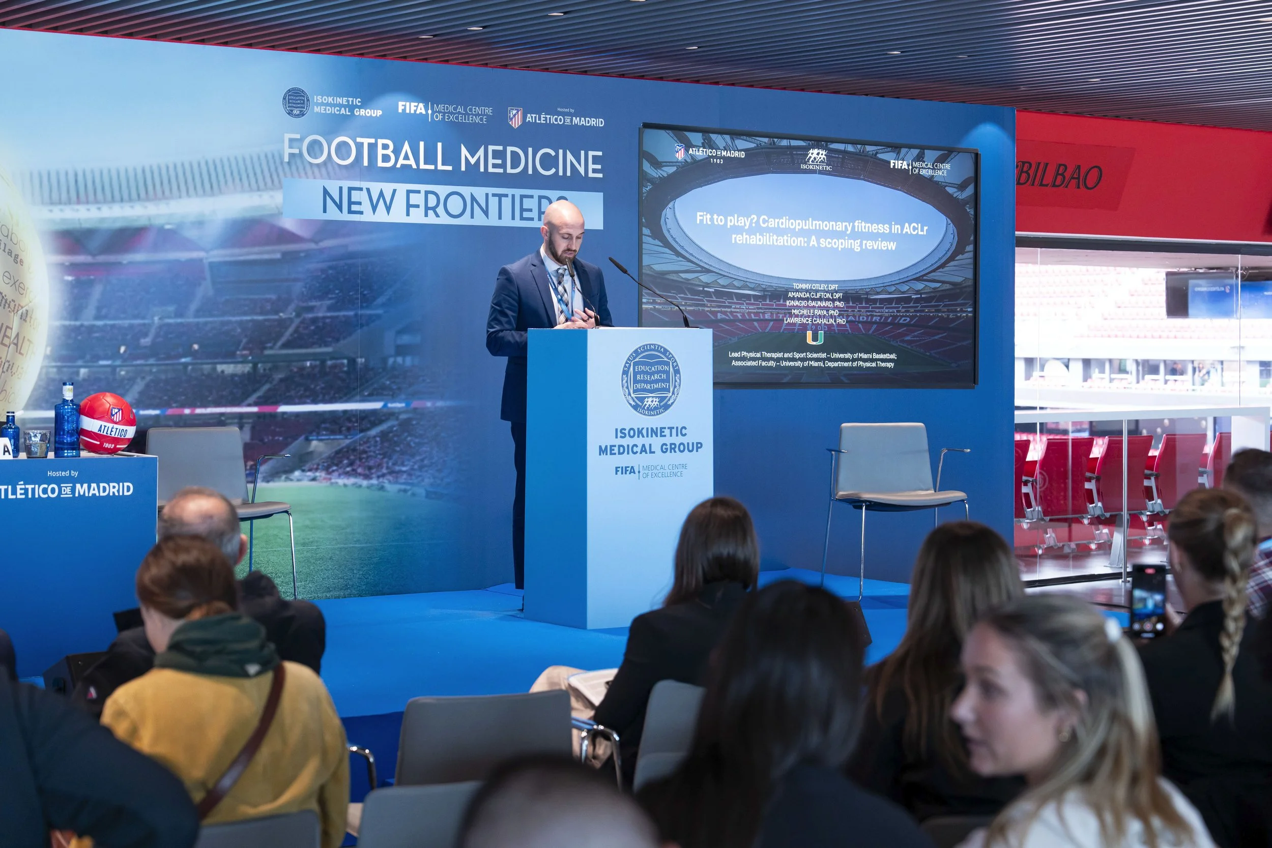 A man in a suit giving a presentation at a conference with a large screen behind him displaying a medical research topic. There are attendees sitting and listening, and a blue panel with logos and text related to football medicine in the background.