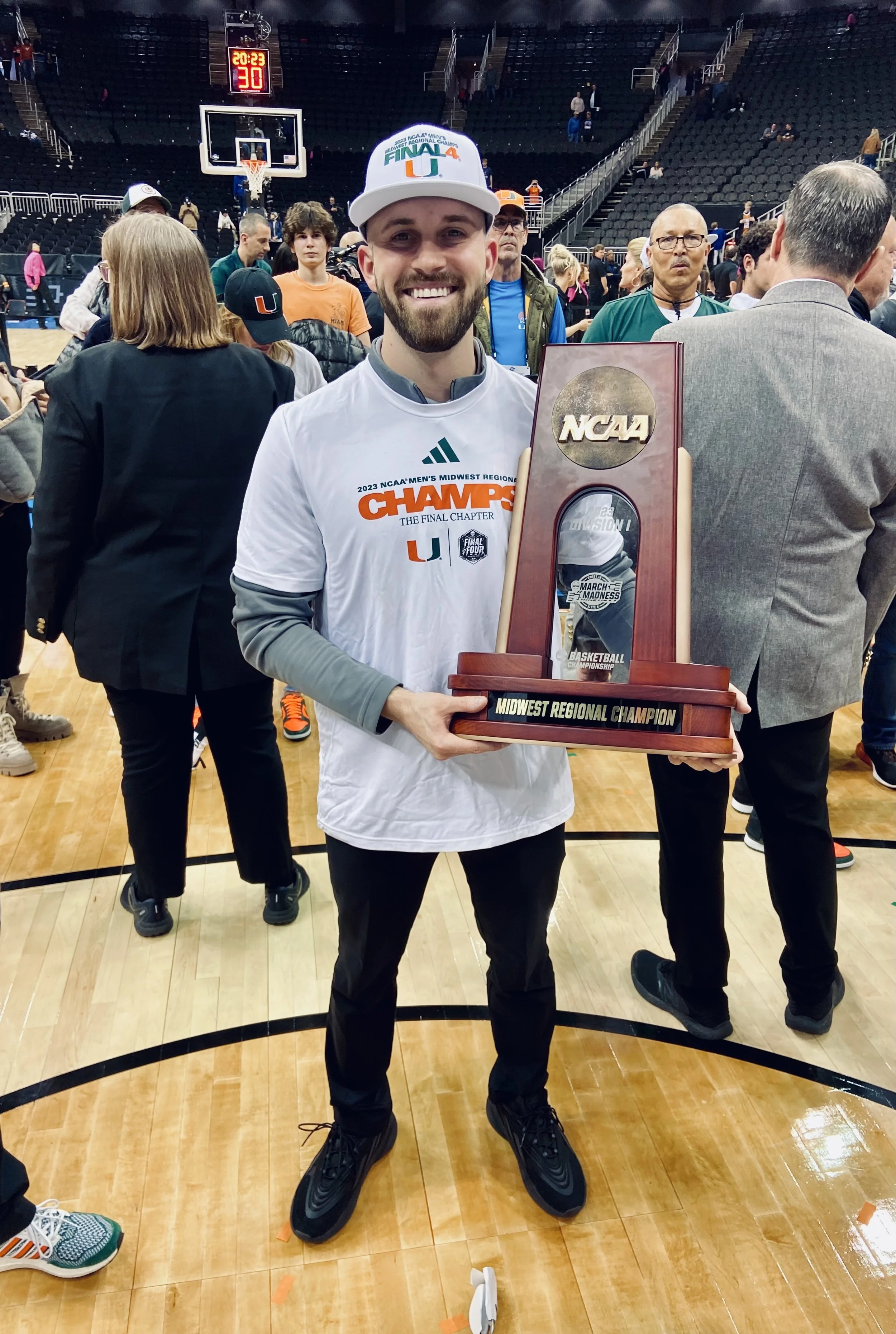 Tommy Otley celebrating  with the Midwest Regional Champion trophy on a basketball court. He is wearing a white t-shirt with event details and a white cap. There are people and a scoreboard in the background, with seats surrounding the court.