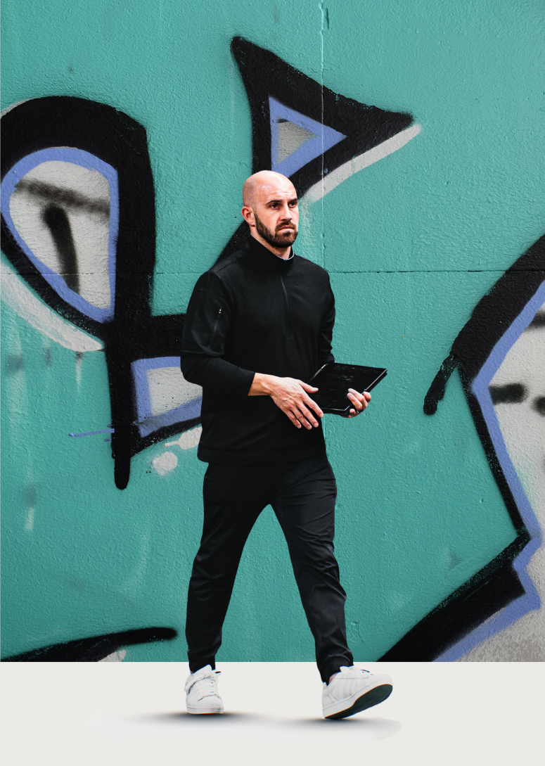 A man with a bald head and beard wearing a black tracksuit and white sneakers holding a tablet while walking in front of a graffiti-covered wall.