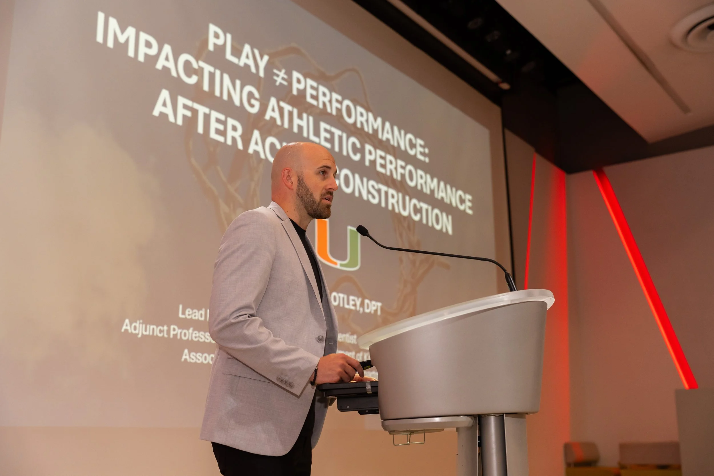 A man in a light gray suit is standing at a podium with a microphone, giving a presentation in a conference room. Behind him, a large screen displays a slide that reads, 'PLAY ≠ PERFORMANCE: IMPACTING ATHLETIC PERFORMANCE AFTER ACL RECONSTRUCTION.'