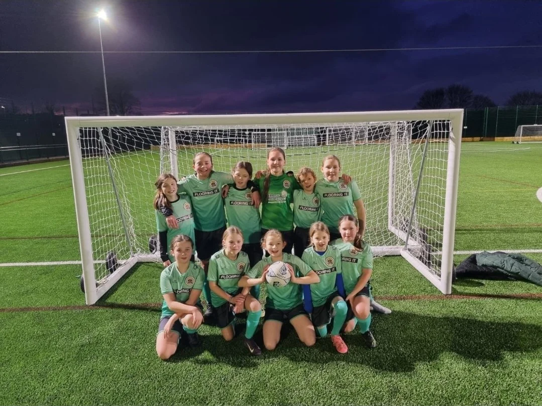 A girls' soccer team of ten players in green uniforms posing in front of a goal on a soccer field at dusk. Five girls kneel in front, holding a soccer ball, while five girls stand behind with their arms around each other's shoulders. The sky is dark 