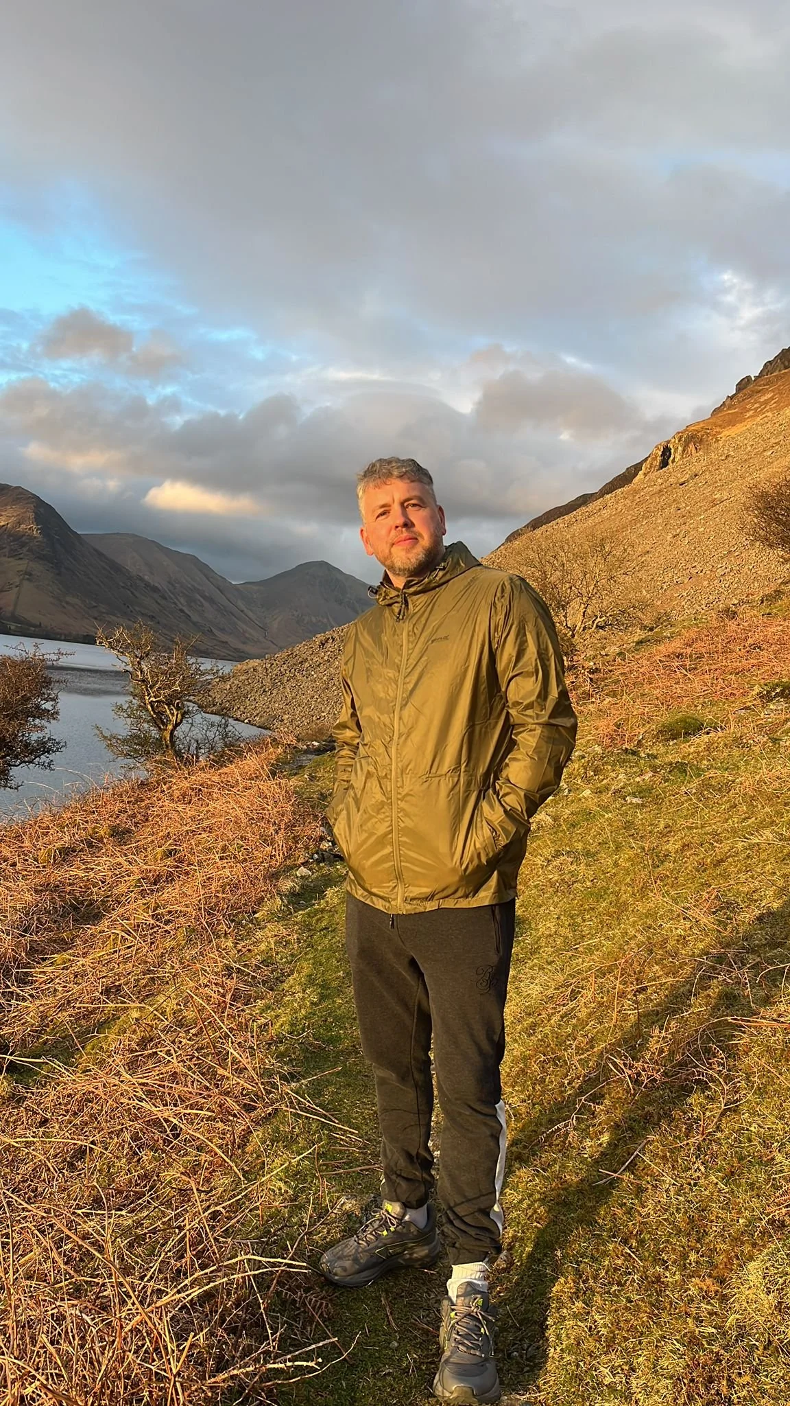 A man dressed in a brown jacket and dark pants standing on a trail beside a body of water, with mountains and cloudy sky in the background during golden hour.