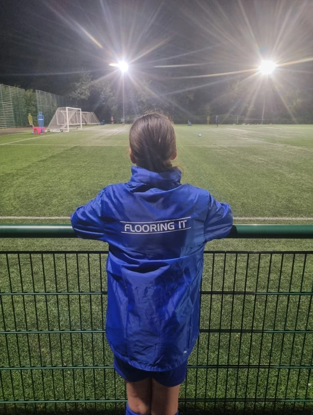 Child in a blue jacket watching a soccer game at night on a well-lit field.