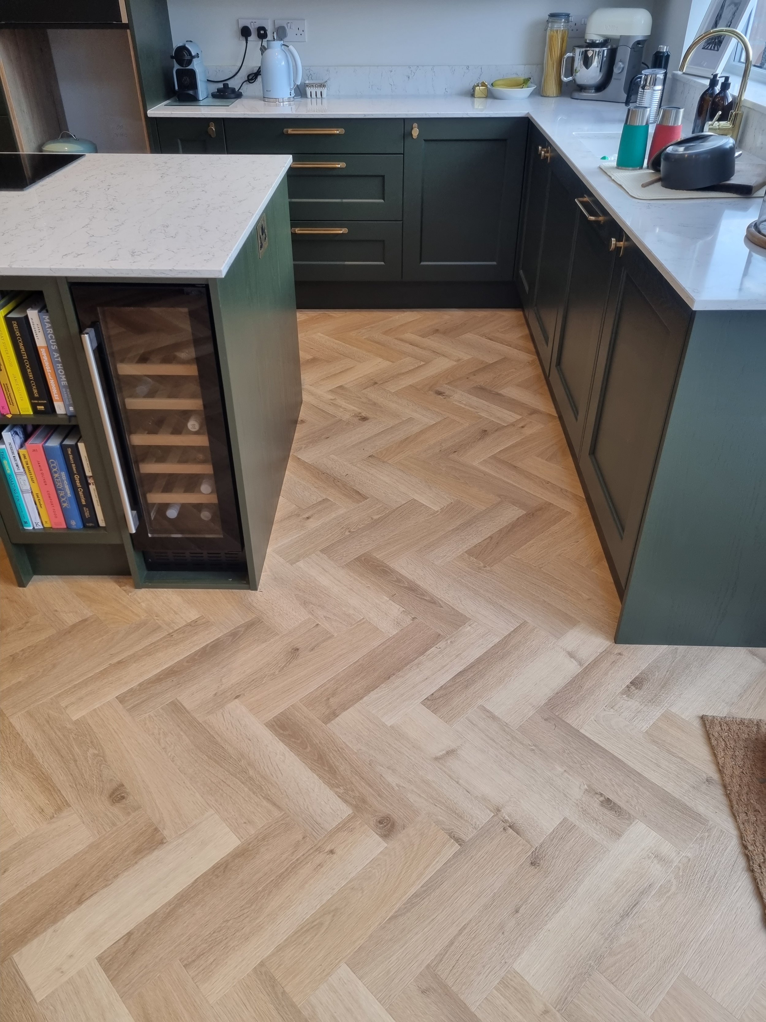 Kitchen with dark green cabinets, white marble countertops, and wooden herringbone-patterned floor. Counter with various appliances and a small wine fridge.