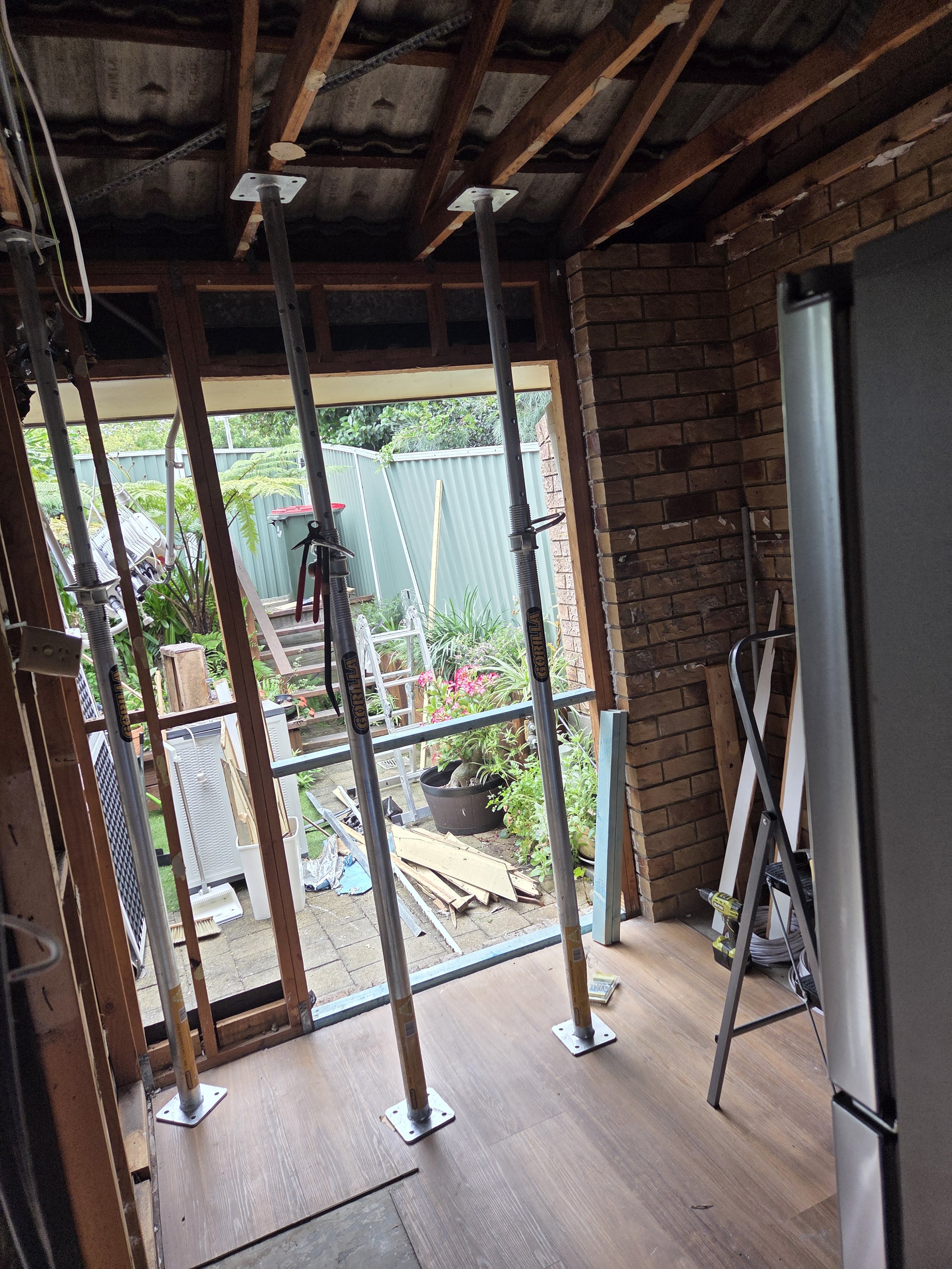 View of a partially renovated interior room with construction tools, roof support metal poles, and a doorway leading outside to a garden with plants and a green fence.