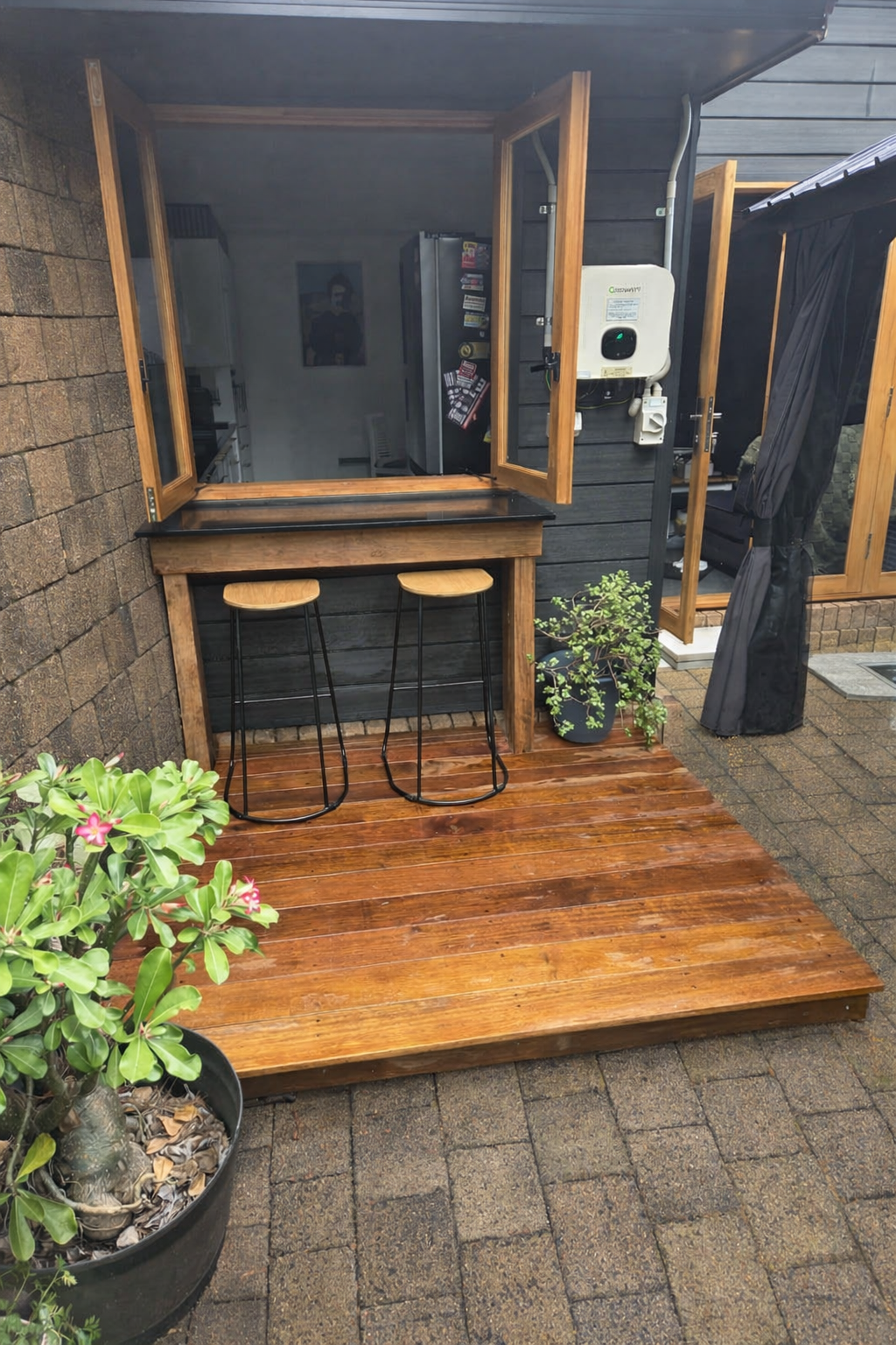 A small outdoor wooden deck with two black barstools, a bar-height wooden table, and potted plants. The deck is attached to a dark gray house with open wooden window shutters and a small indoor kitchen visible inside.