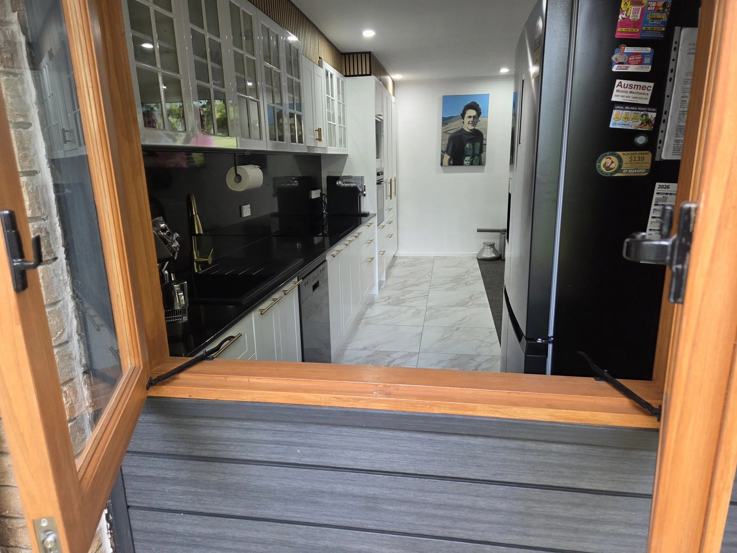 View of a modern kitchen through a wooden-framed french windows with glass panels. The kitchen has white cabinets, black counter tops, a gold faucet, and a floating paper towel holder. A large view of the newly installed kitchen's