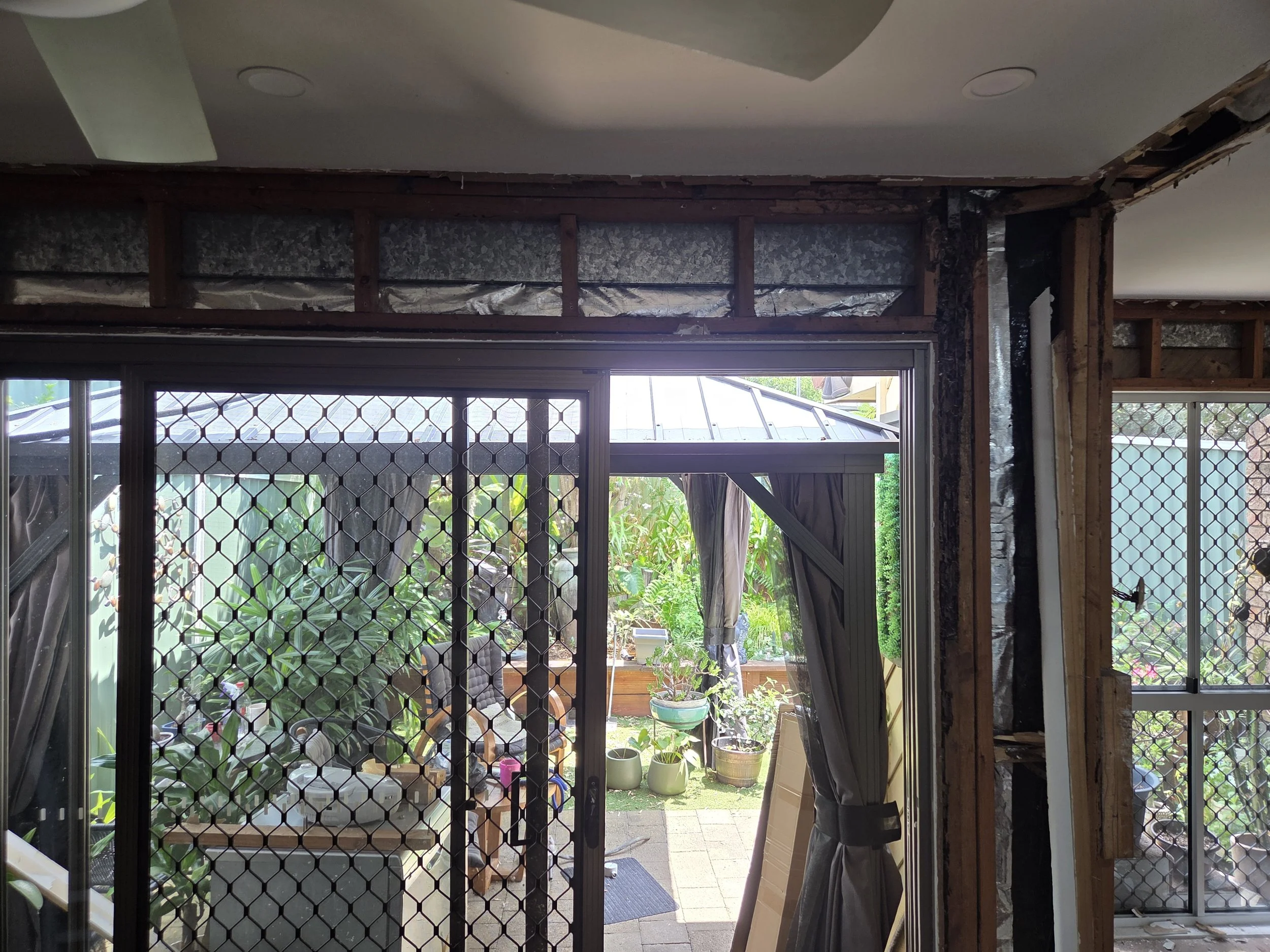 View through a glass door with a diamond-shaped wire security screen, showing potted plants, a patio, and a garden with trees and plants outside.