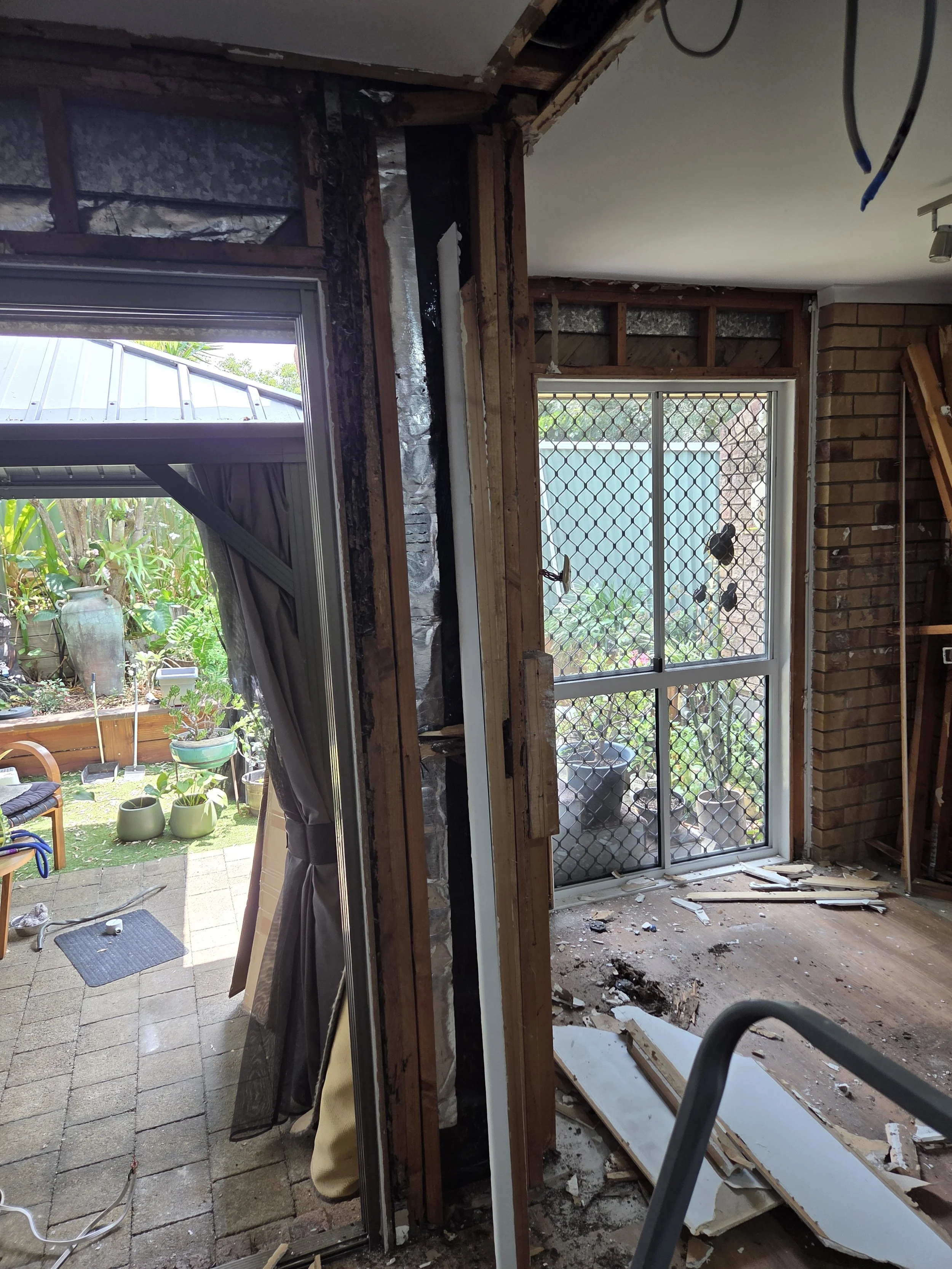 Partial view of a home renovation, showing an open wall with exposed termite damaged framing, insulation, and a sliding glass door with a security screen, leading to a patio garden with potted plants and outdoor furniture.