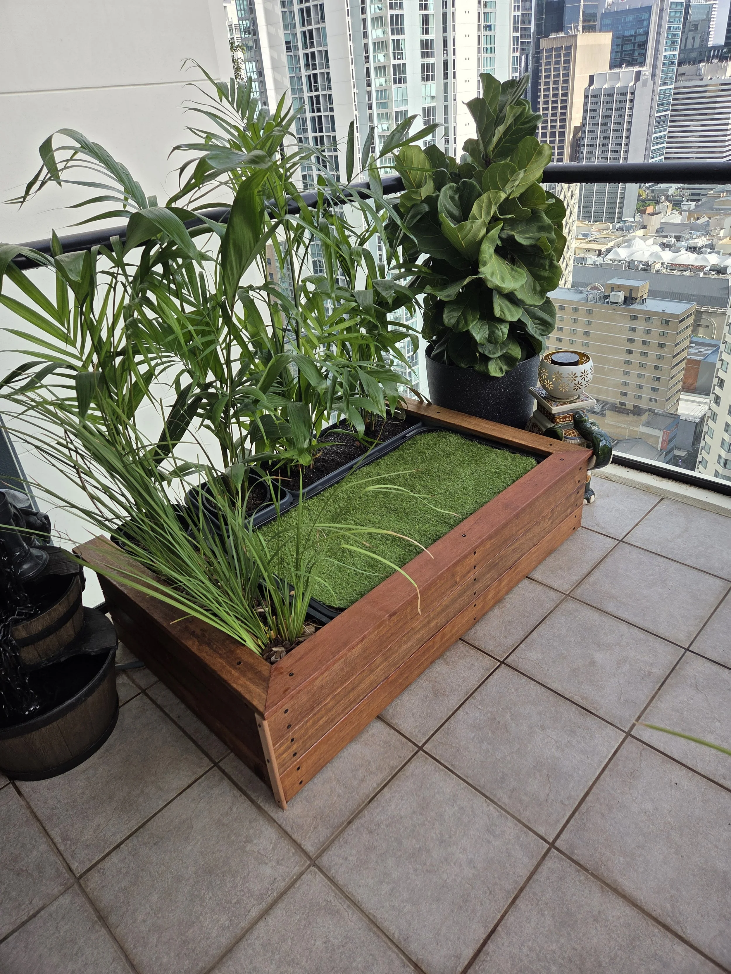 A balcony with potted green plants in a wooden planter, overlooking a city with tall buildings.