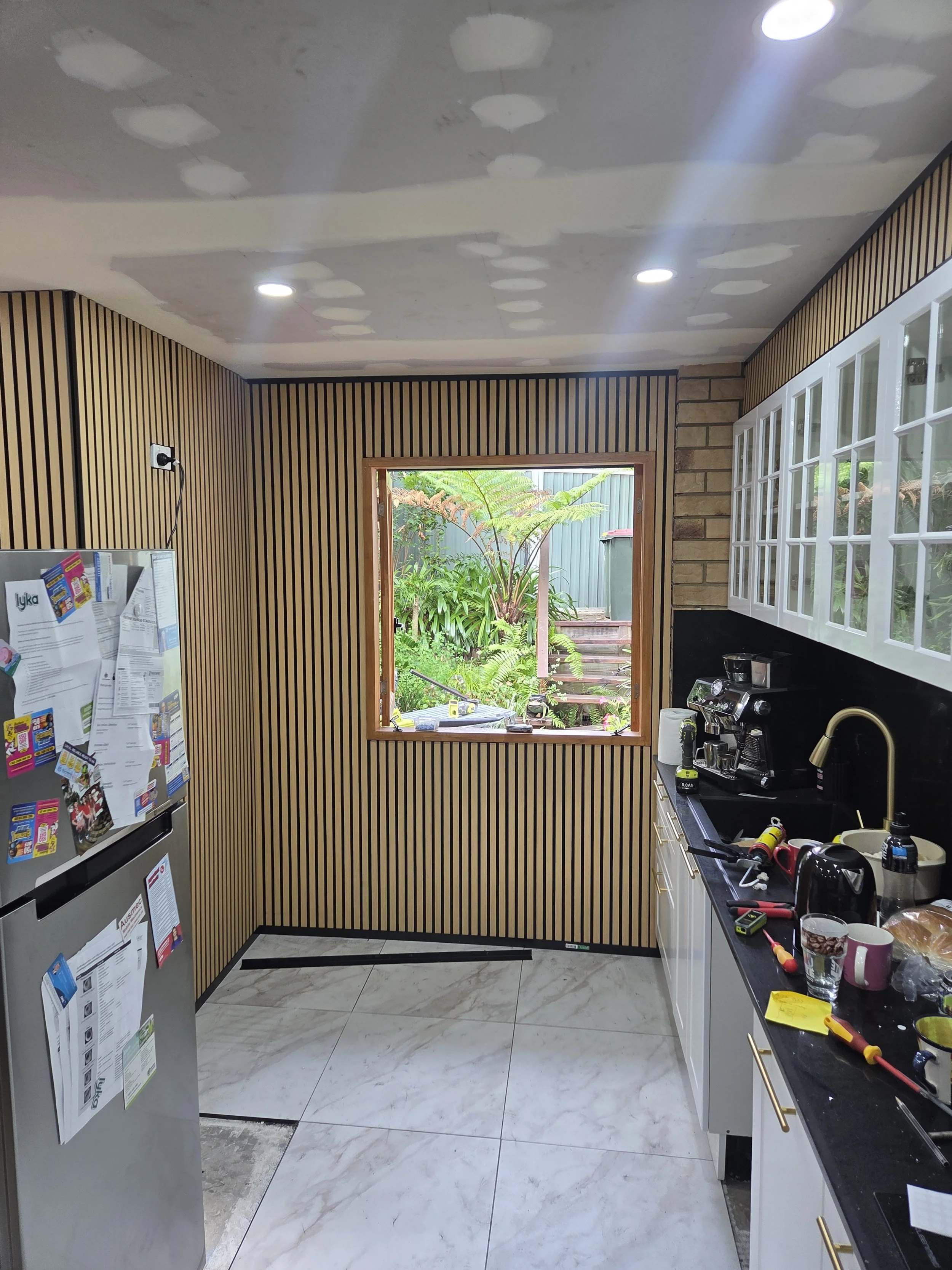 Kitchen with partially finished walls and ceiling, black stone countertops, white cabinets with glass doors, window opening to greenery outside, and a silver refrigerator covered with papers and posters.