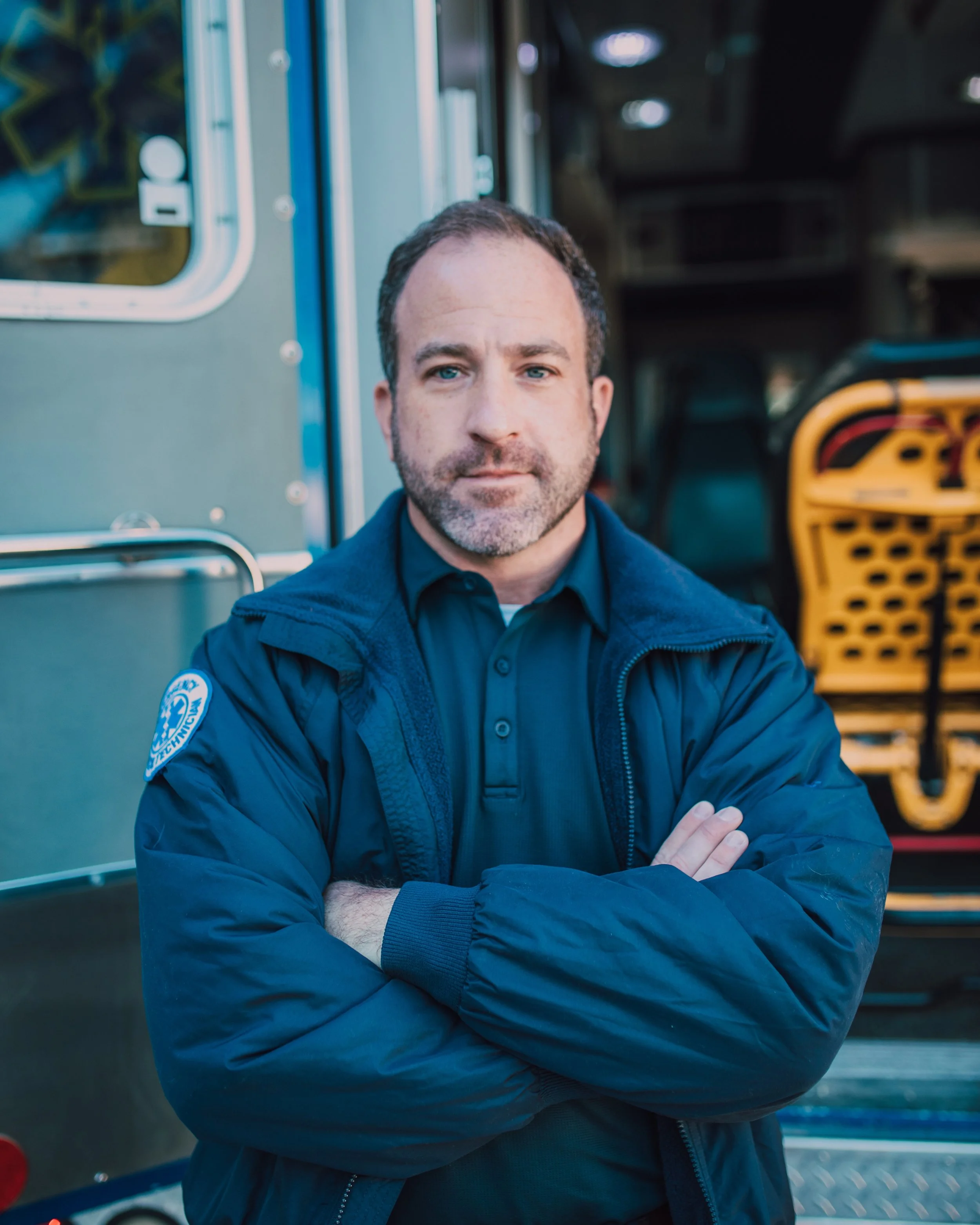 A man with short dark hair, beard, and blue eyes wearing a dark blue uniform with a police badge patch on his left arm, standing with arms crossed in front of emergency vehicles, including an ambulance and a fire truck.