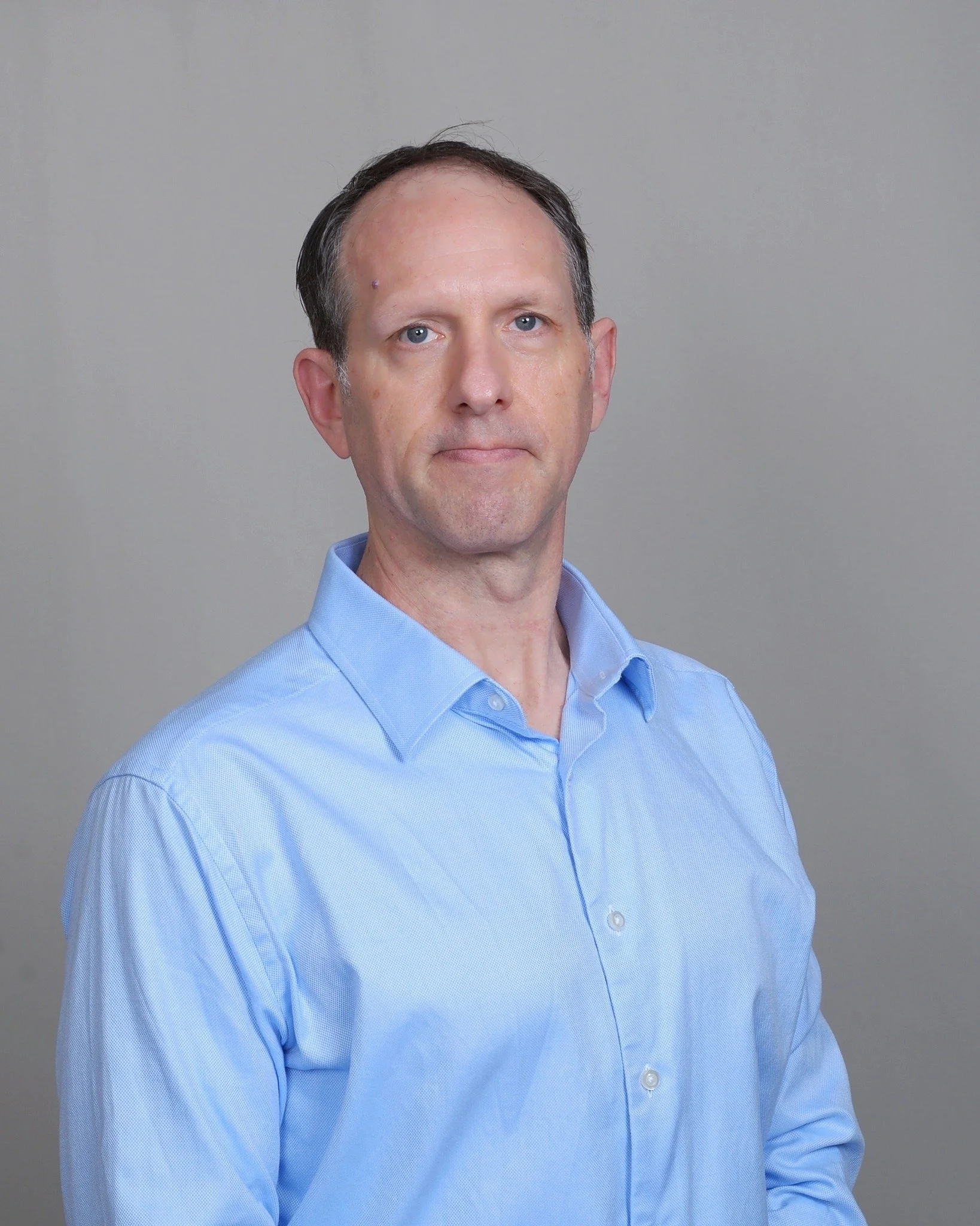 Portrait of a middle-aged man with light skin, blue eyes, and short dark hair with some gray, wearing a blue dress shirt, against a plain gray background.