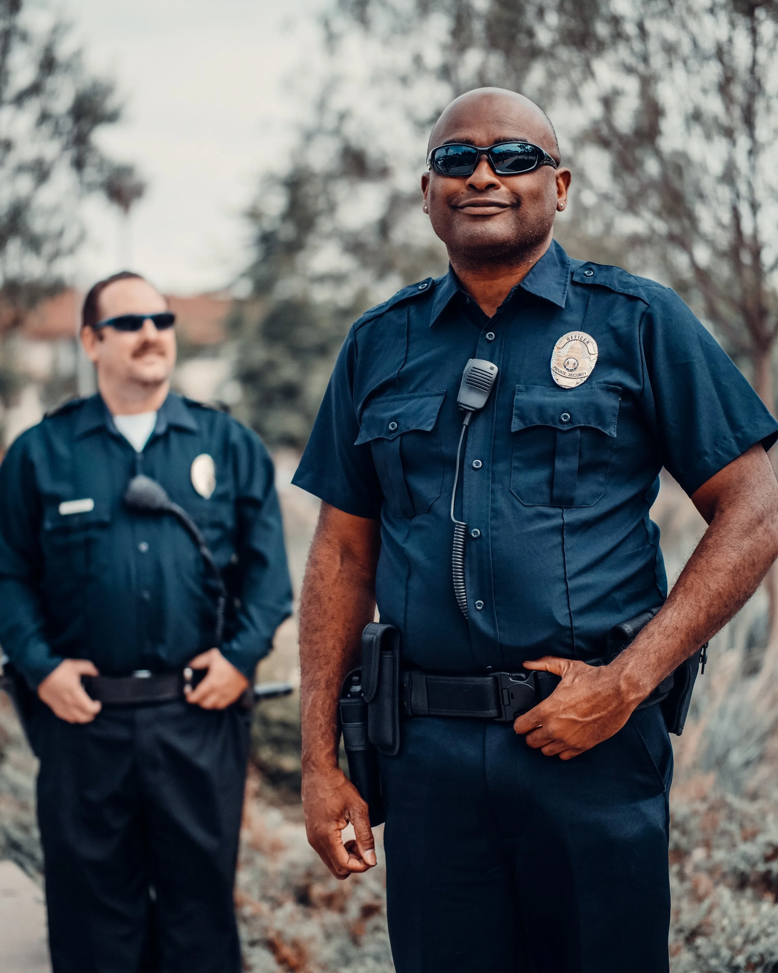 Two police officers standing outdoors with trees in the background. The officer in the foreground is an African American man wearing sunglasses, a police uniform, and a badge. The officer in the background is a Caucasian man also wearing sunglasses and a police uniform.