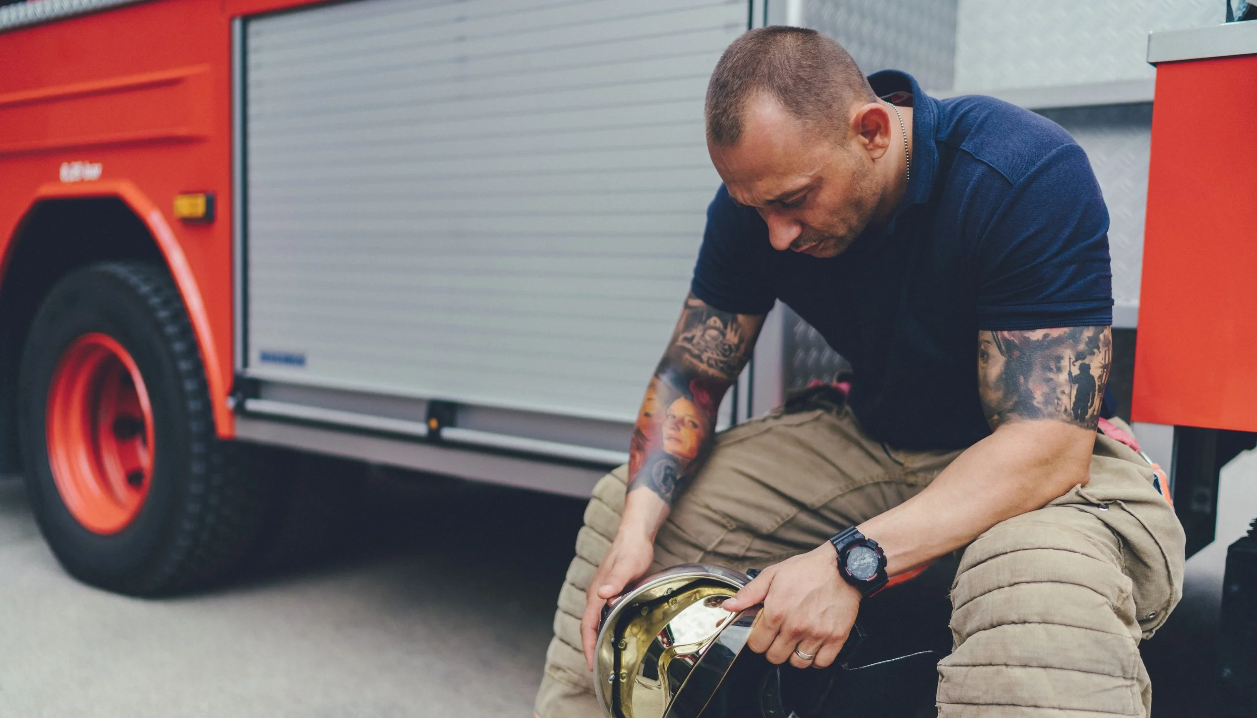 A firefighter kneeling on the ground, holding a firefighter helmet, with a fire truck in the background.