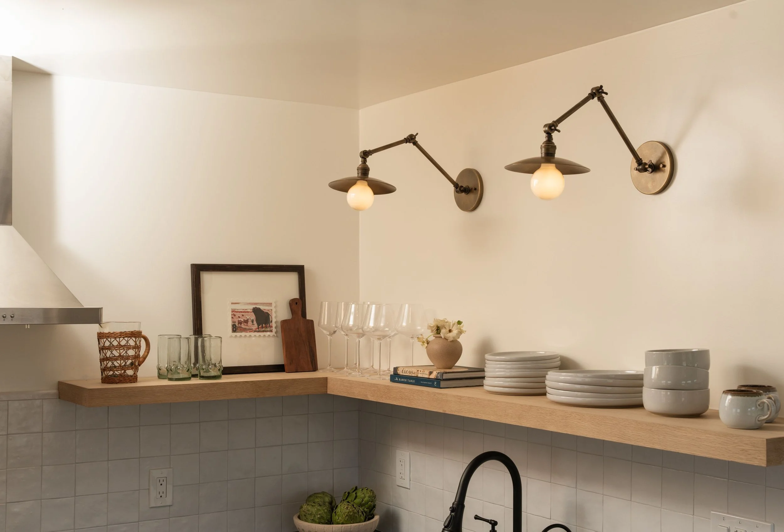 Kitchen with a wooden shelf holding glassware, bowls, and a small vase, two wall-mounted lamps with exposed bulbs above, framed picture on the wall, and green artichokes in a bowl near the sink.