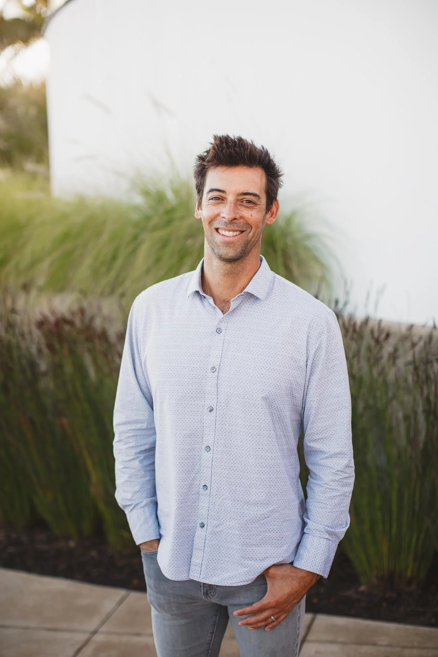 A man with dark hair and a light beard, wearing a white, button-down shirt and gray pants, standing outdoors in front of tall grass and a white wall, smiling at the camera.