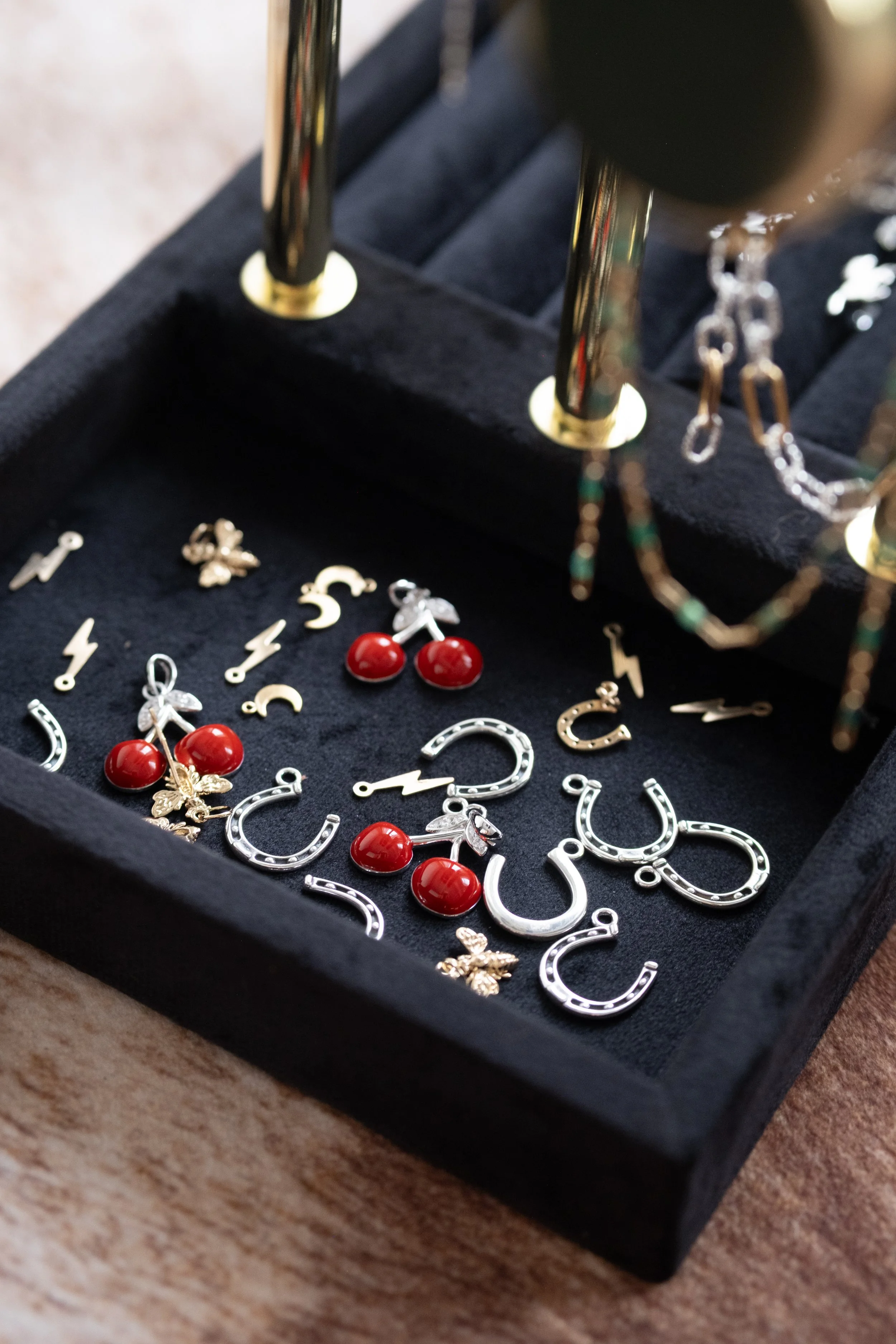 Close-up of a black jewelry display tray with silver horseshoe charms, red cherry charms, and small silver charms shaped like a key, butterfly, and lightning bolt. Some jewelry is hanging from metal rods above the tray, and a chain is visible on the 