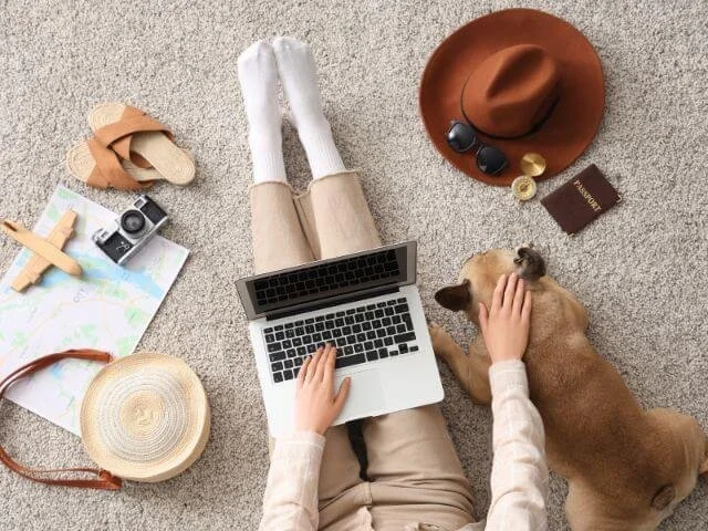 Person using a laptop on the floor surrounded by travel items and a dog lying beside them.
